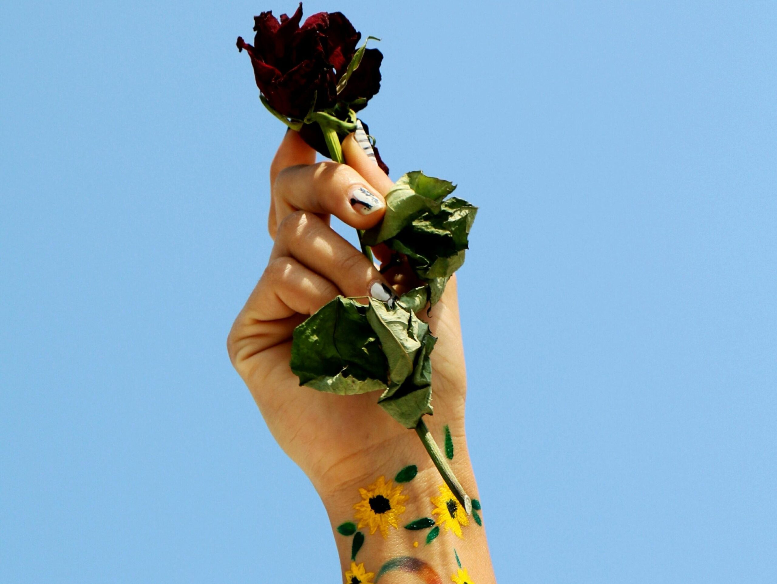 A hand raised against a clear blue sky holding a wilted red rose. The wrist and forearm are painted with bright yellow sunflowers and green leaves, symbolizing resilience and growth.