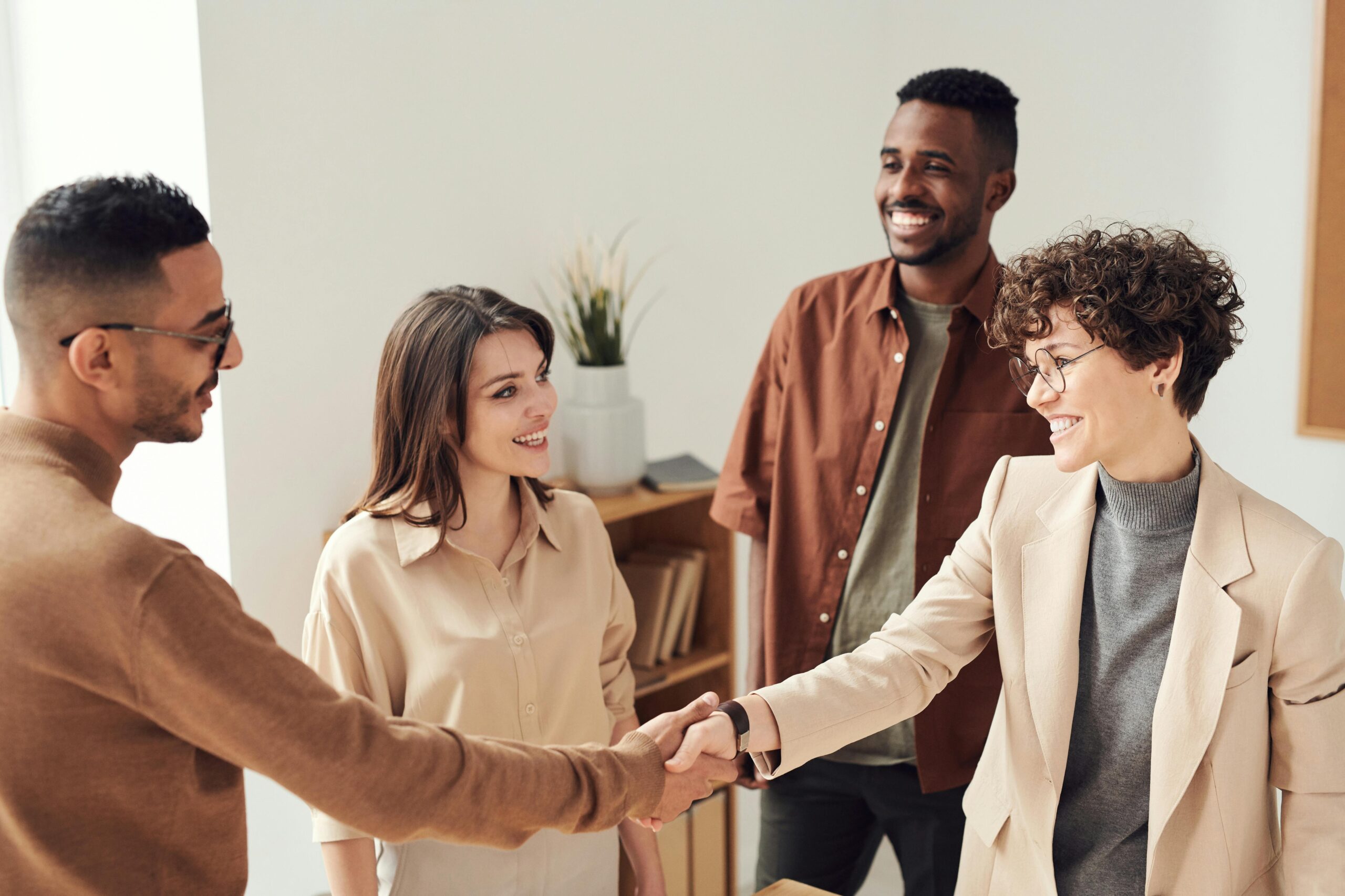 Four colleagues in an office smiling together, with two of them shaking hands while the others look on, symbolizing connection and trust