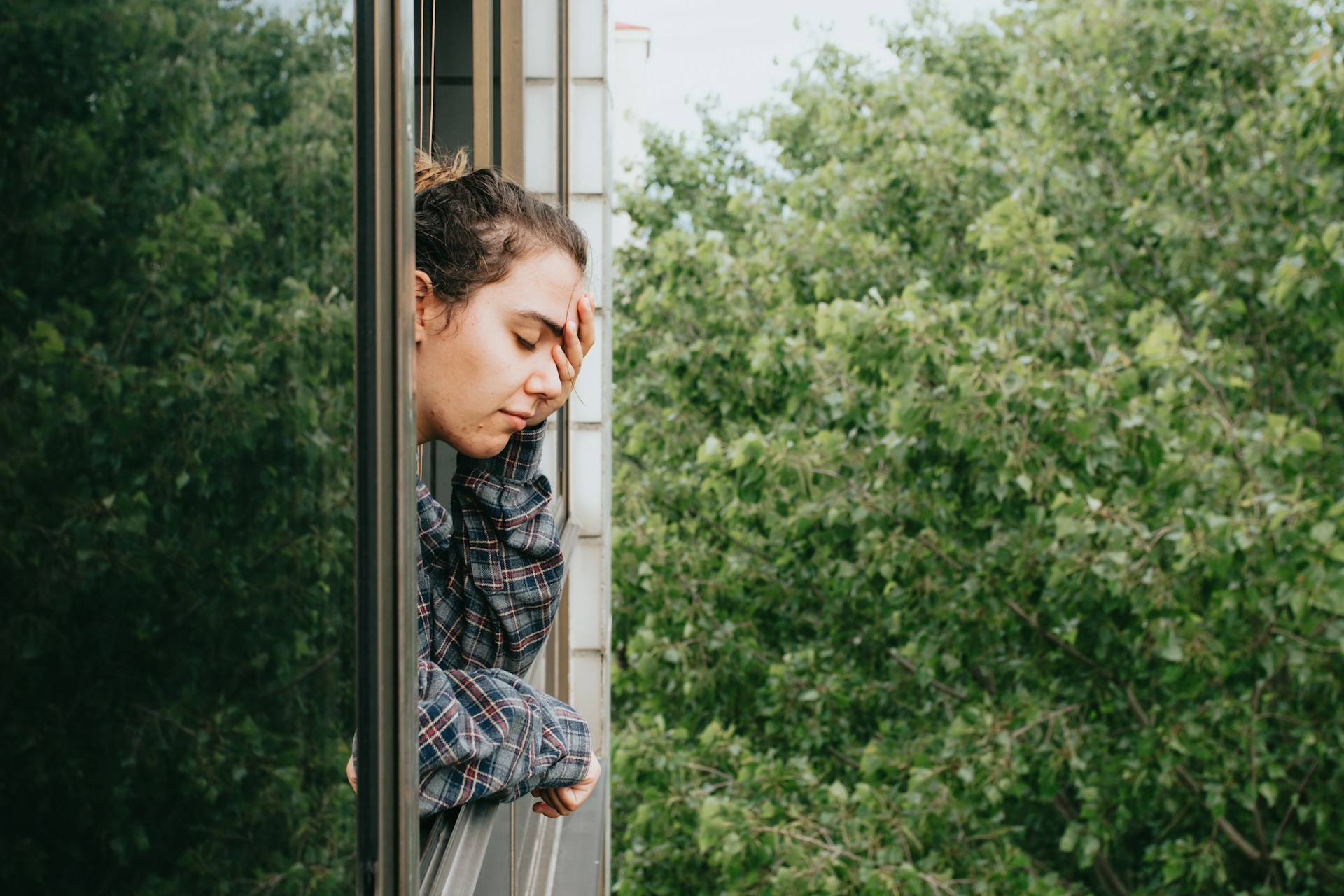 A woman gazes thoughtfully out of a sunlit window, watching the bright summer day unfold outside, reflecting the theme of mindfulness and coping with seasonal stress.