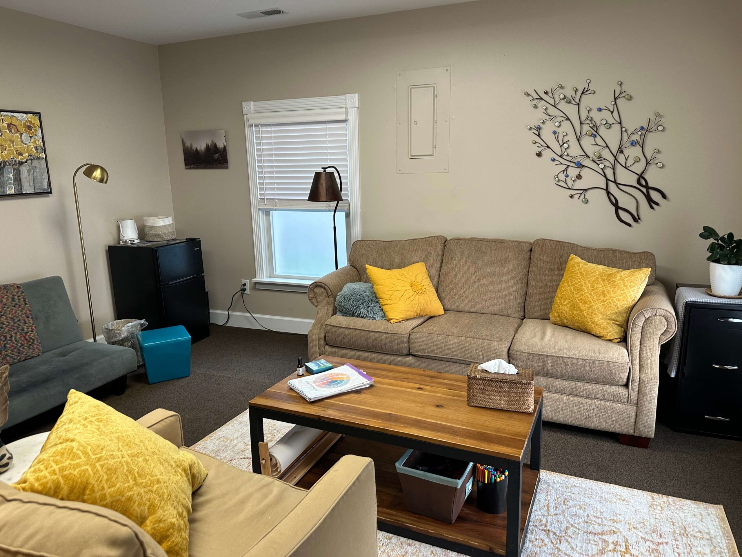 A cozy therapy room with a tan couch, yellow pillows, and a rustic wood coffee table at the Joy Spring Mental Health office in Boone, NC.