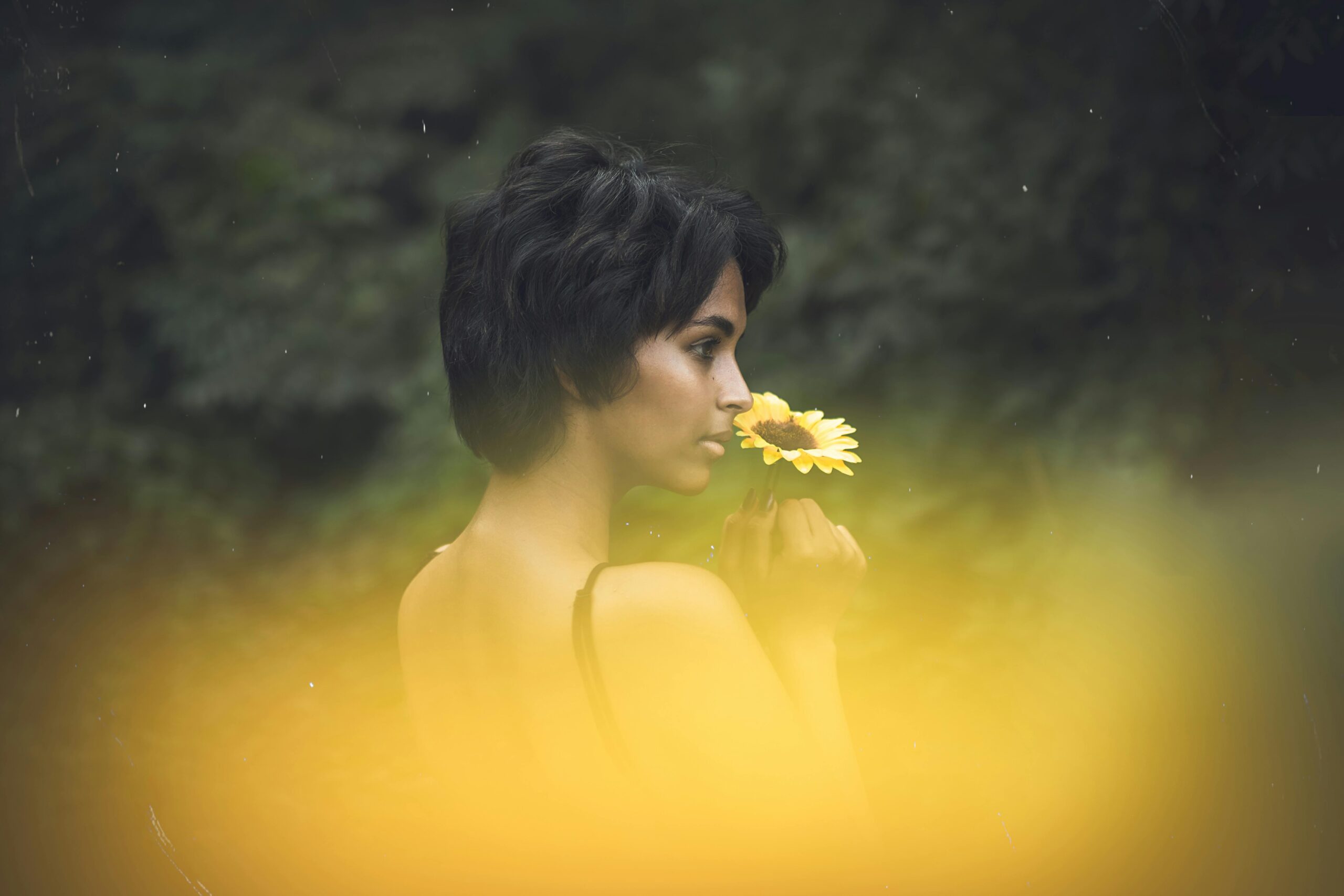 Woman holding a sunflower near her face, standing in soft golden light with a blurred natural background.
