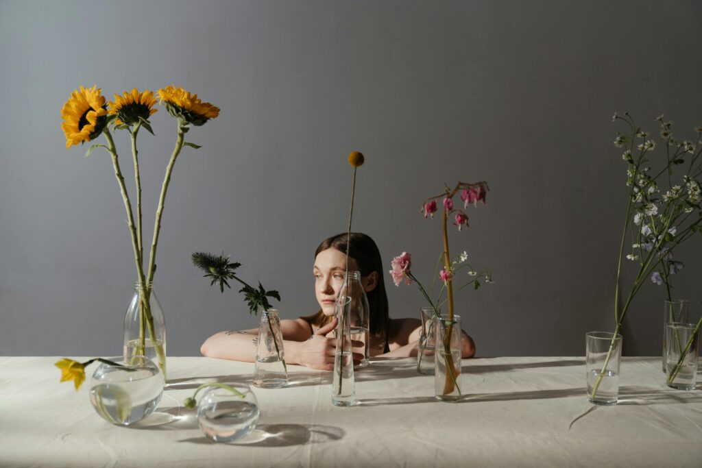 "A contemplative woman sits behind sparse vases of flowers, visually capturing the balance between idle habits and quiet survival in Lazy Habits: The Survival Side.