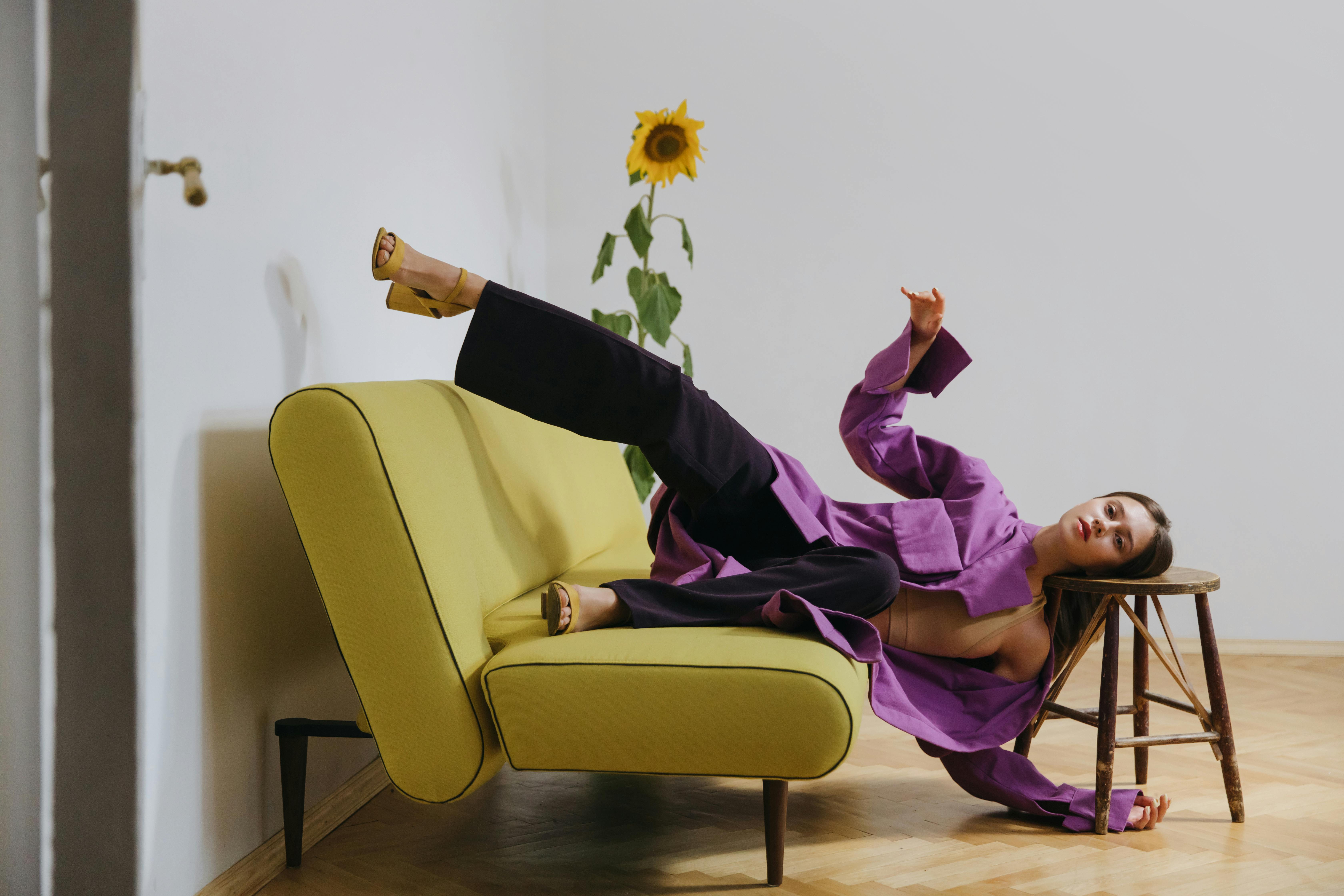A woman in a flowing purple blouse and black pants holds an offbeat, almost precarious pose across a yellow sofa and wooden stool. Her lifted leg and resting head create a tension between imbalance and control, symbolizing the delicate harmony of body and mind. A tall sunflower in the background adds a quiet sense of grounding.