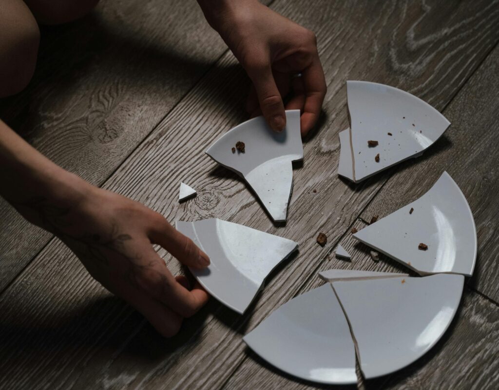 Close-up of hands gathering broken white ceramic plate pieces scattered across a wooden floor, symbolizing the process of mending or reintegration.
