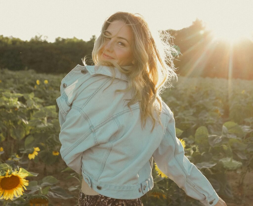 A woman in a light denim jacket stands in a sunflower field at sunset, turning back with a gentle smile as golden sunlight filters through her hair.