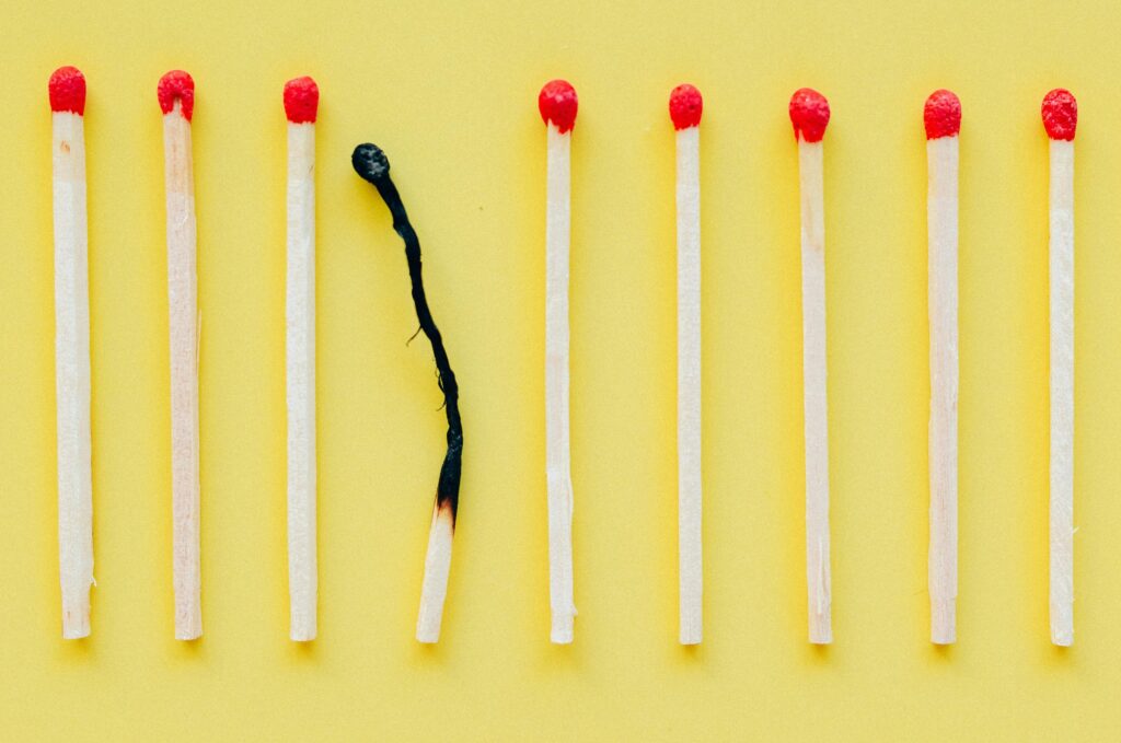 A row of matches on a yellow background, with one burnt-out match in the middle among unlit matches, symbolizing burnout, meaning, and mental health.