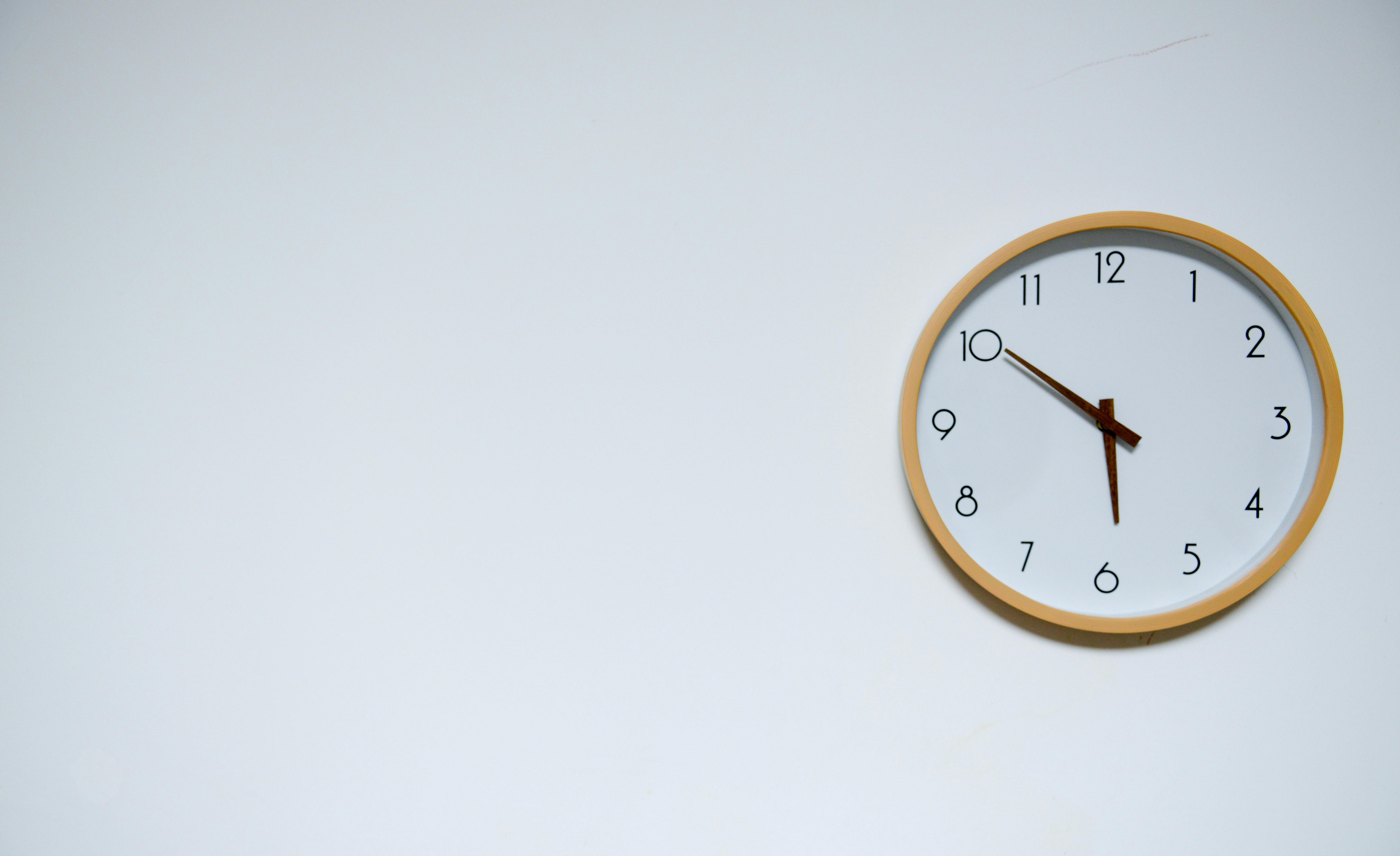 A minimalist wall clock with a wooden frame shows the time as 10:50 against a blank white wall, symbolizing the quiet pressure of passing time and the feeling that time is never enough.