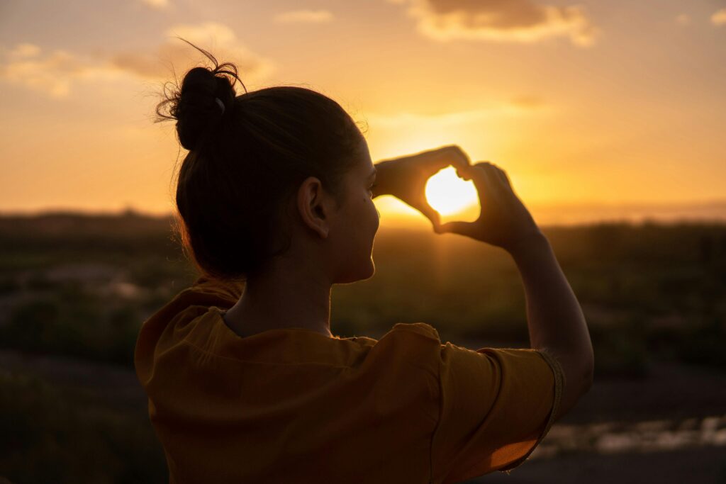 A woman stands at sunset, forming a heart shape with her hands around the glowing sun, symbolizing love, hope, and the journey of healing.