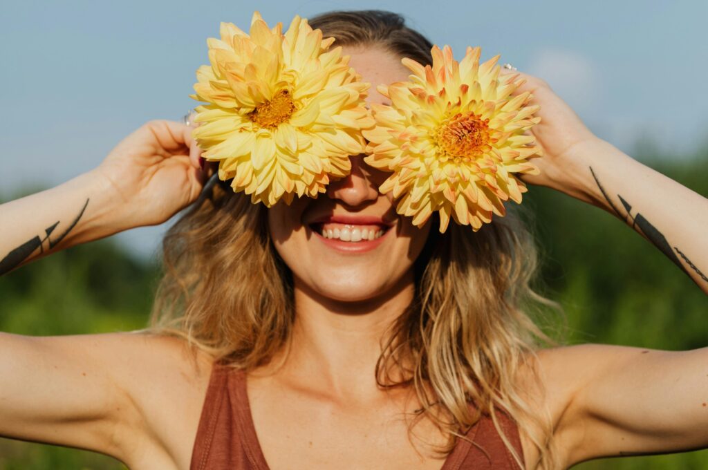 Smiling woman holding two large yellow flowers over her eyes on a sunny day, enjoying nature and expressing joy and mindfulness.