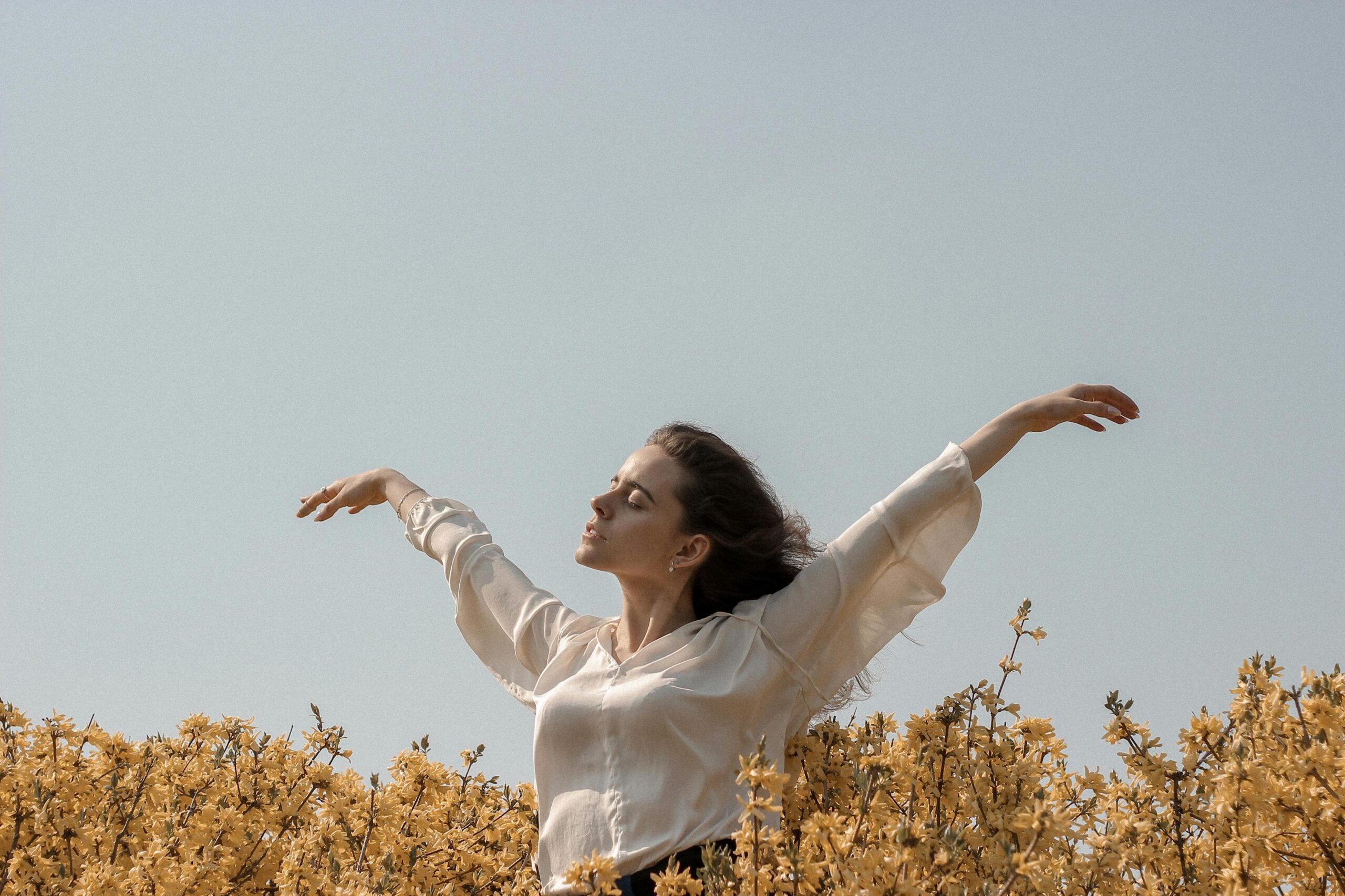 A woman stands in a field of yellow flowers under a clear sky, eyes closed and arms outstretched, embracing the sunlight with a peaceful, freeing expression.