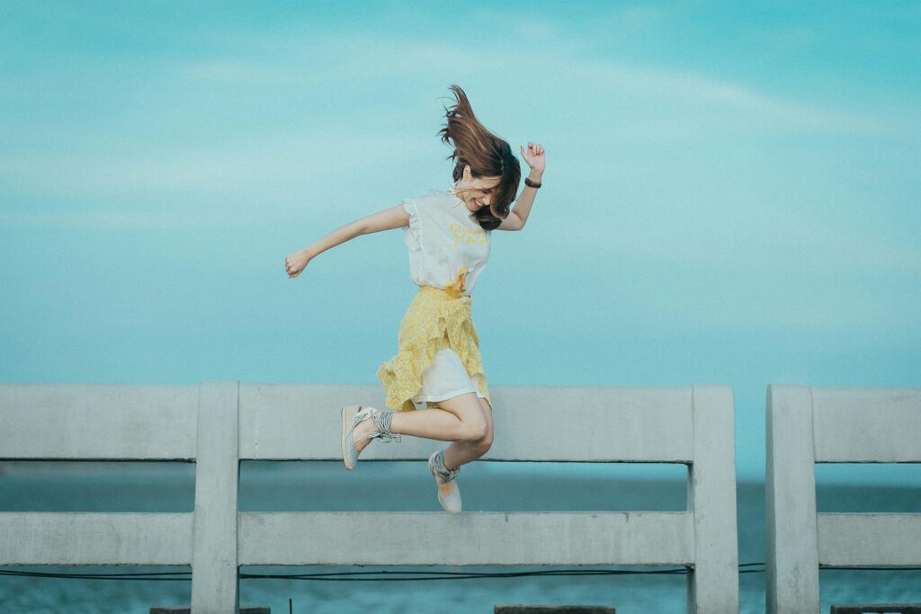 A young woman joyfully jumping in the air on a seaside bridge under a clear blue sky, wearing a yellow skirt and white top — symbolizing freedom, happiness, and emotional wellness.