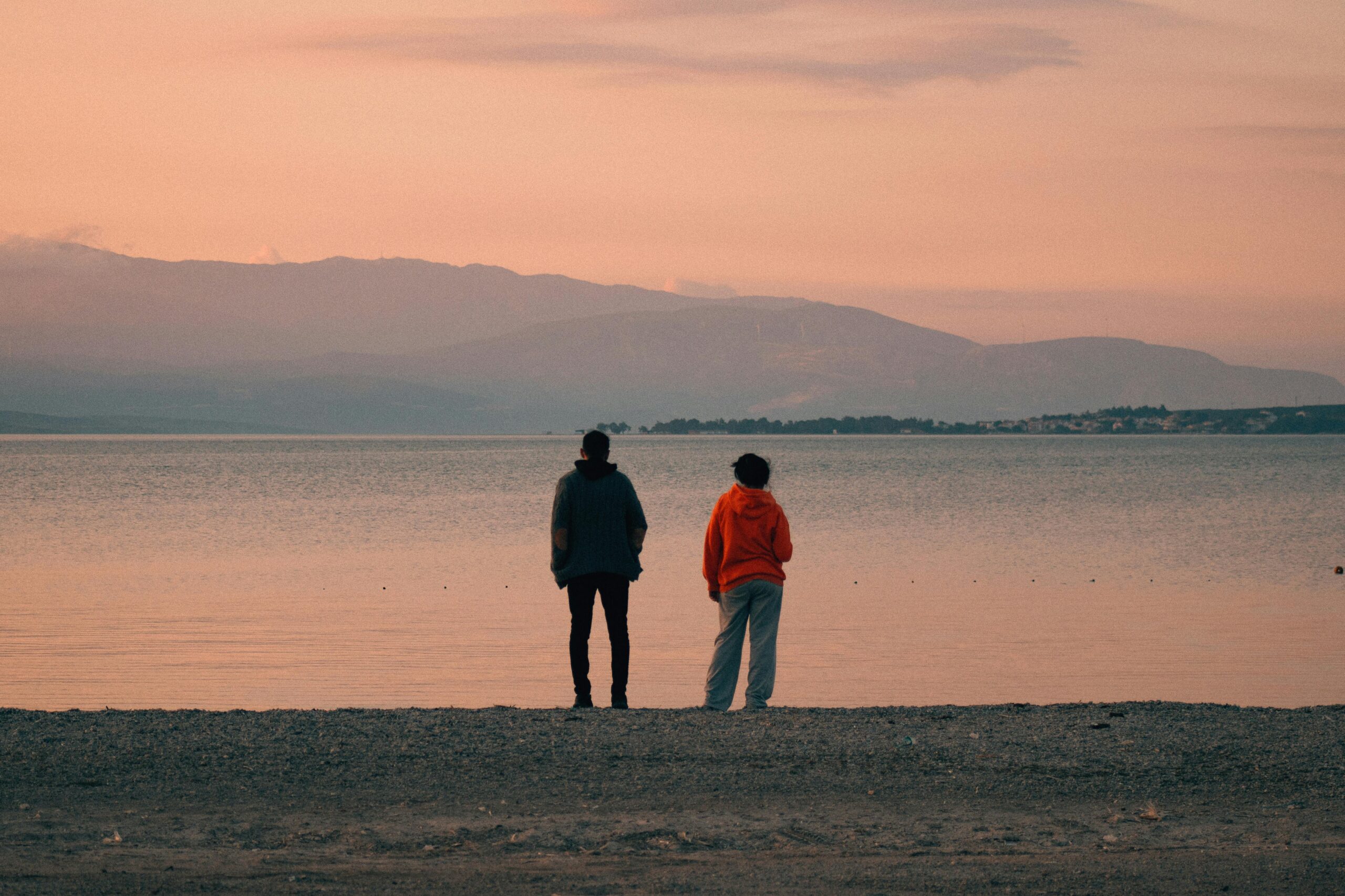 Two people stand quietly on a pebbled shore, gazing across a calm lake toward distant mountains under a pink-orange sunset sky. One person wears a gray hoodie and dark pants, the other a bright orange hoodie and light sweatpants. The peaceful scene conveys stillness, reflection, and the quiet comfort of companionship.