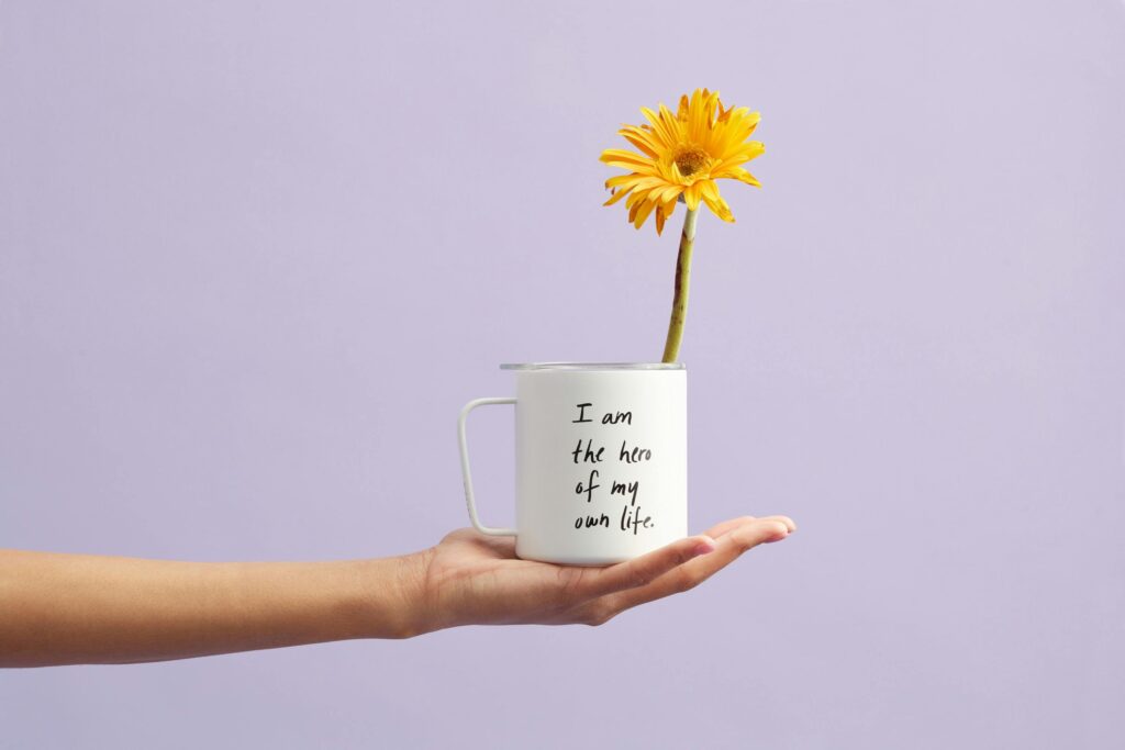 A hand gently holds a white mug with the words “I am the hero of my own life” written on it. A single bright yellow flower blooms from the mug against a soft lavender background, symbolizing growth, self-empowerment, and calm transformation.