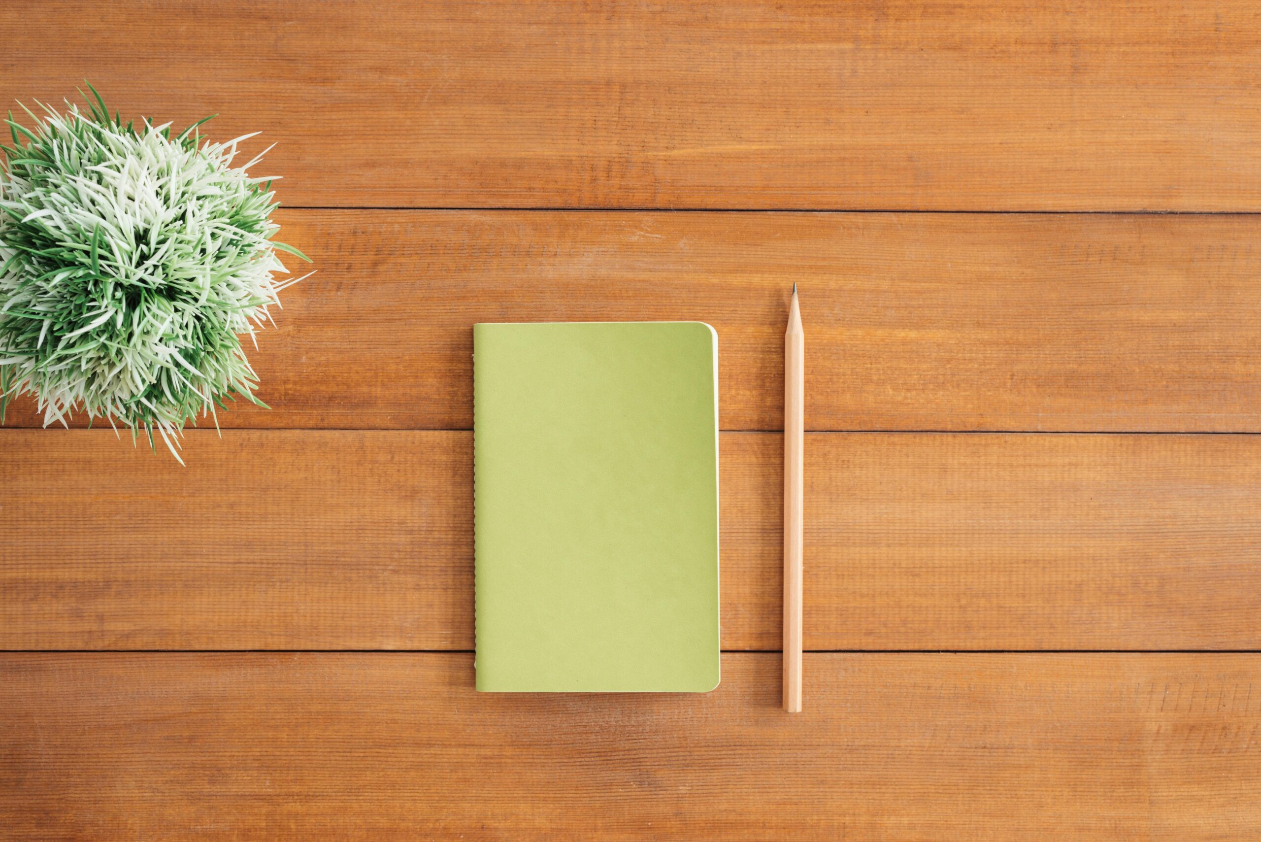 A minimalist flat lay featuring a yellow notebook and a wooden pencil neatly placed on a warm wooden desk, with a small decorative plant to the side, symbolizing reflection, growth, and new beginnings.