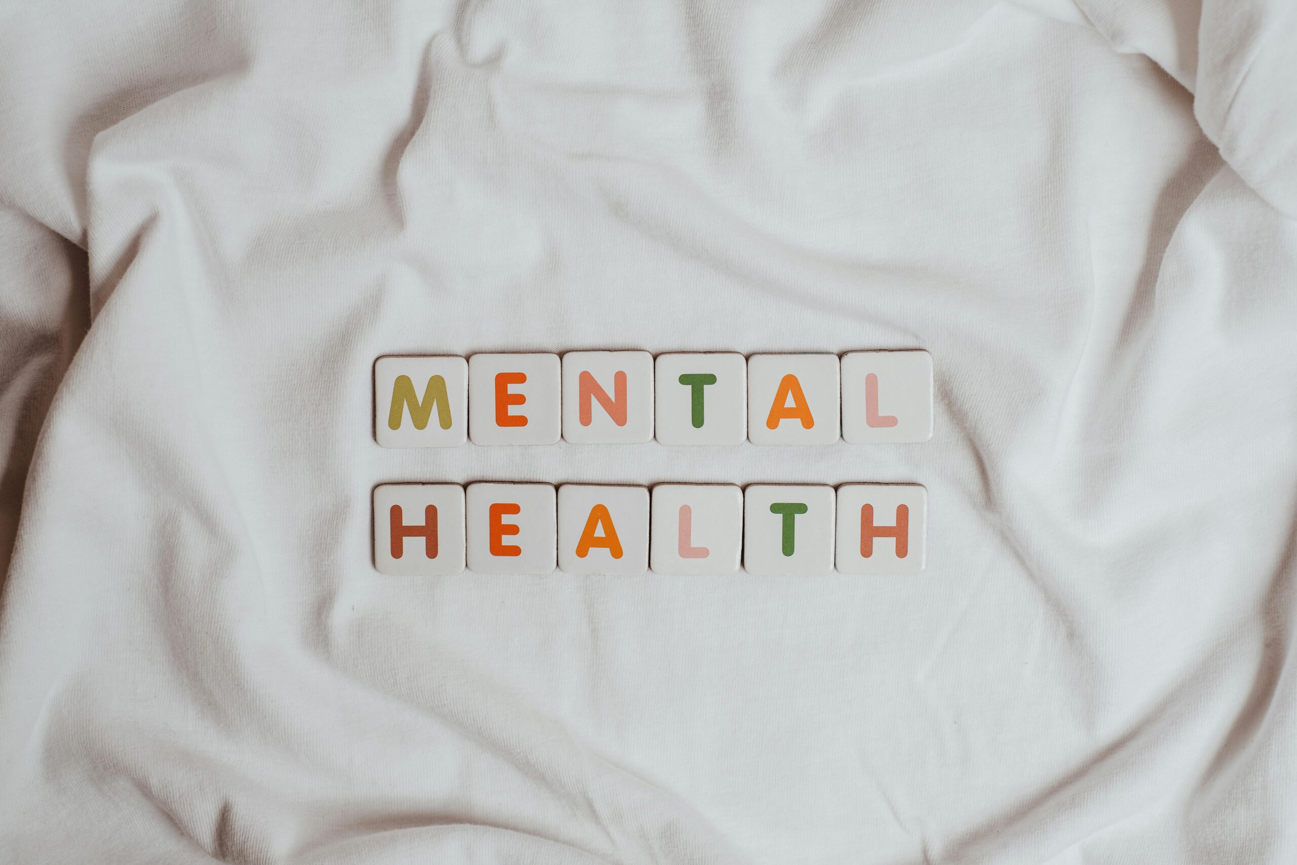 Colorful letter tiles spelling out 'MENTAL HEALTH' arranged neatly on a soft white fabric background, symbolizing calmness, reflection, and awareness for mental well-being.
