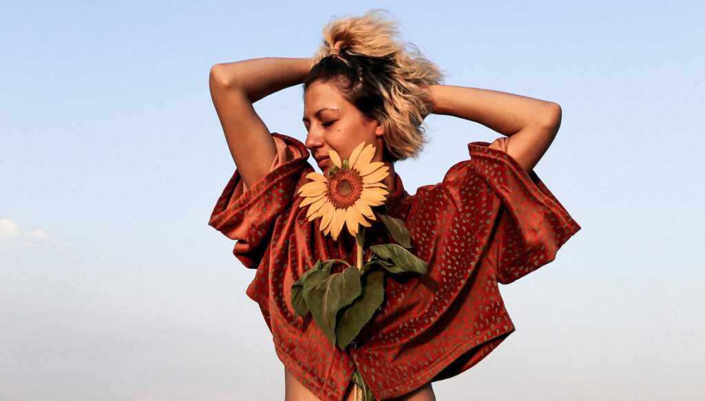 A woman stands under a clear blue sky, eyes closed and smiling softly, holding a sunflower close to her chest. She wears a flowing rust-colored patterned top with her arms raised behind her head, radiating confidence and self-acceptance.