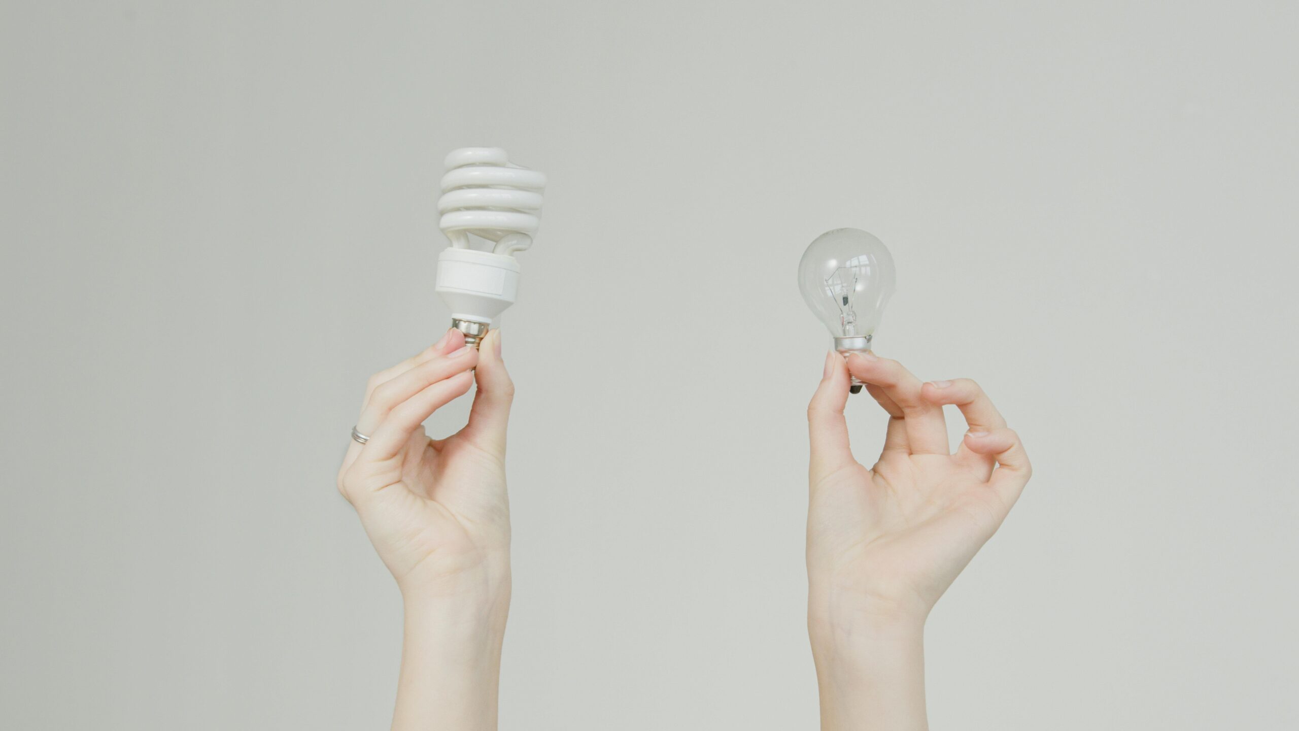 Two hands holding different light bulbs—a modern energy-efficient spiral bulb and a traditional clear incandescent bulb—symbolizing comparison and choice.