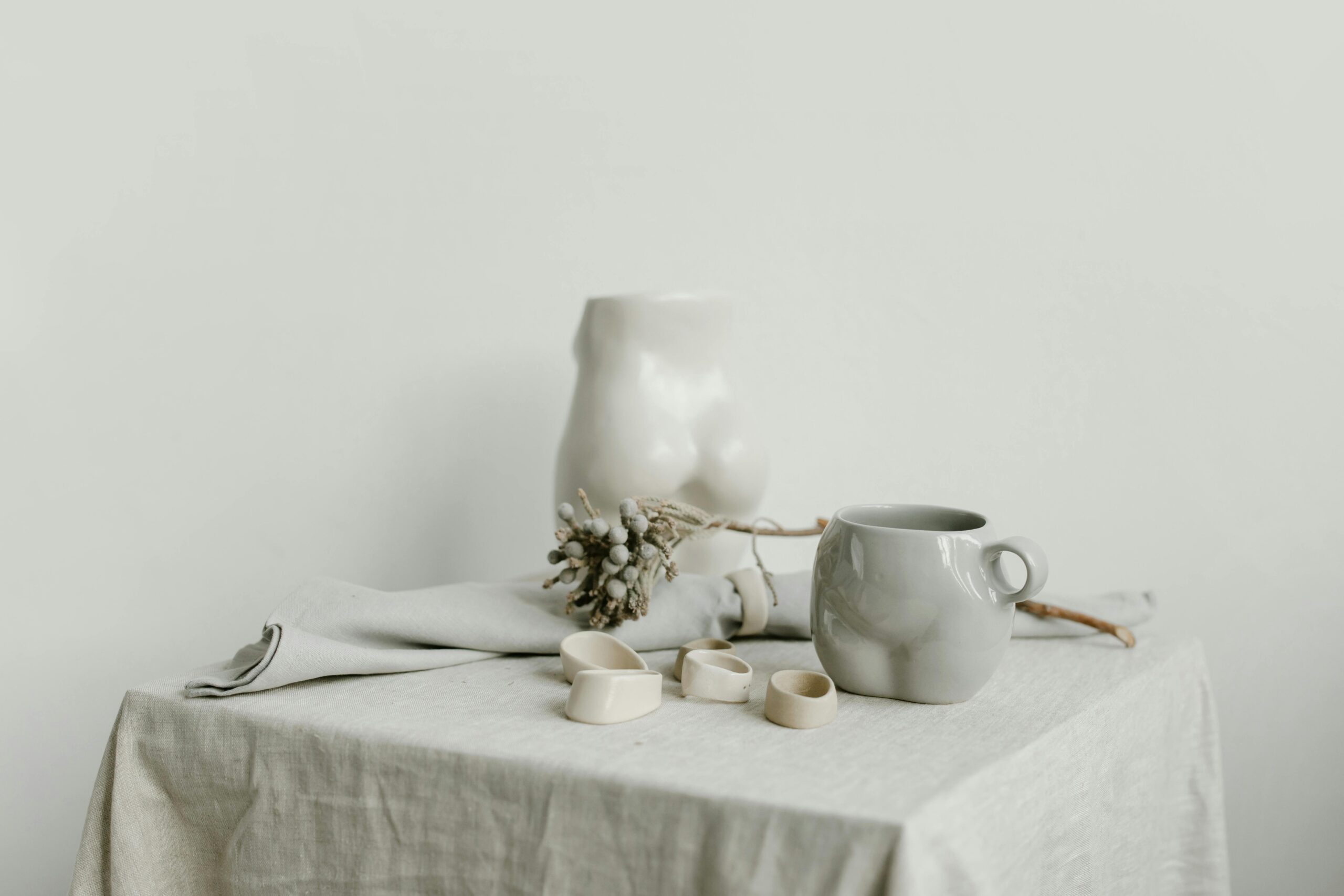 A minimal, neutral-toned still life featuring a grey ceramic mug, small cream-colored ceramic pieces, a cloth napkin, and a sculptural torso-shaped vase on a linen-covered table, styled with a dried botanical stem.