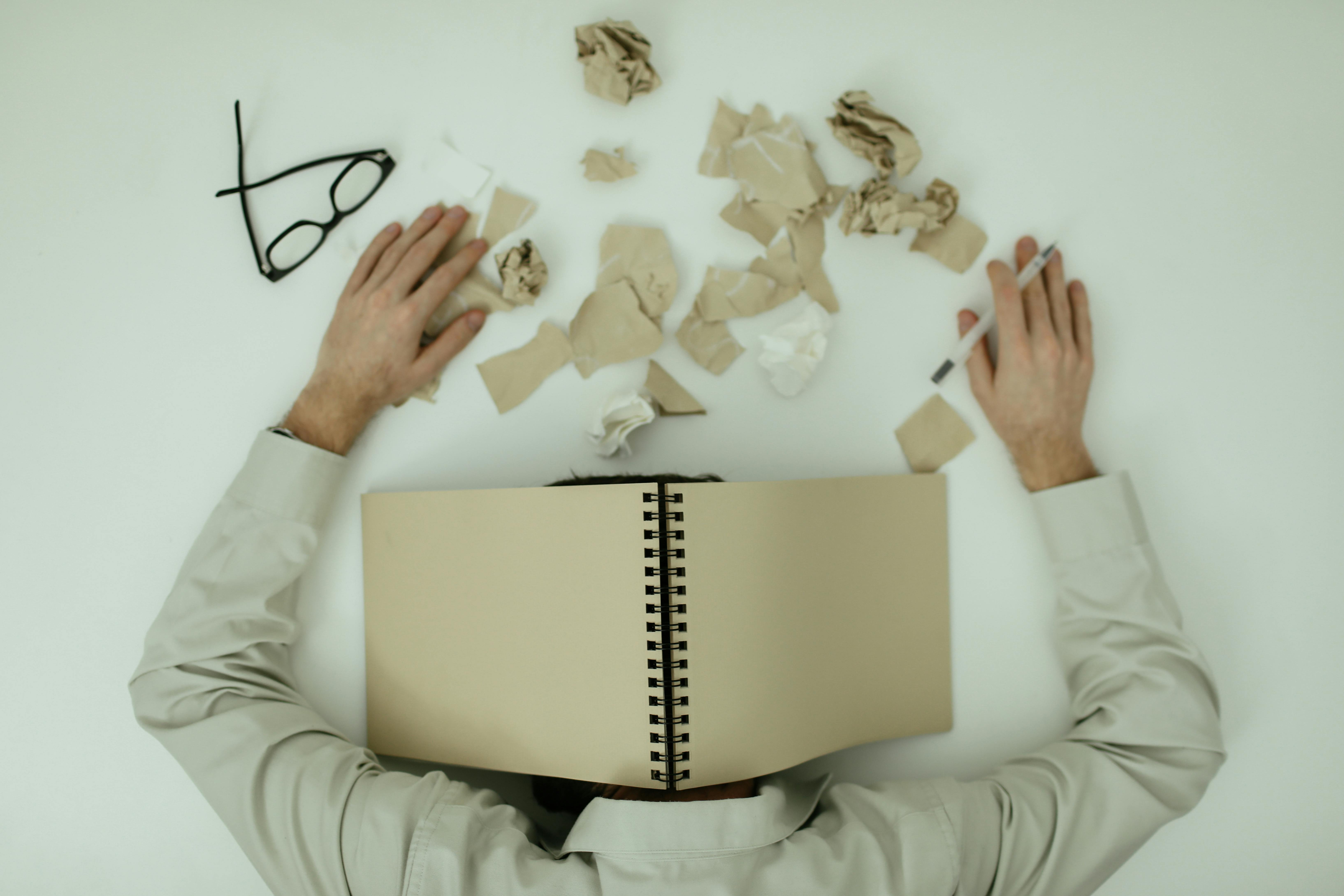 A person wearing a light-colored shirt lies face-down on a desk with an open notebook covering their head, surrounded by crumpled paper, a pen, and eyeglasses—visually representing burnout, overwhelm, and creative exhaustion.