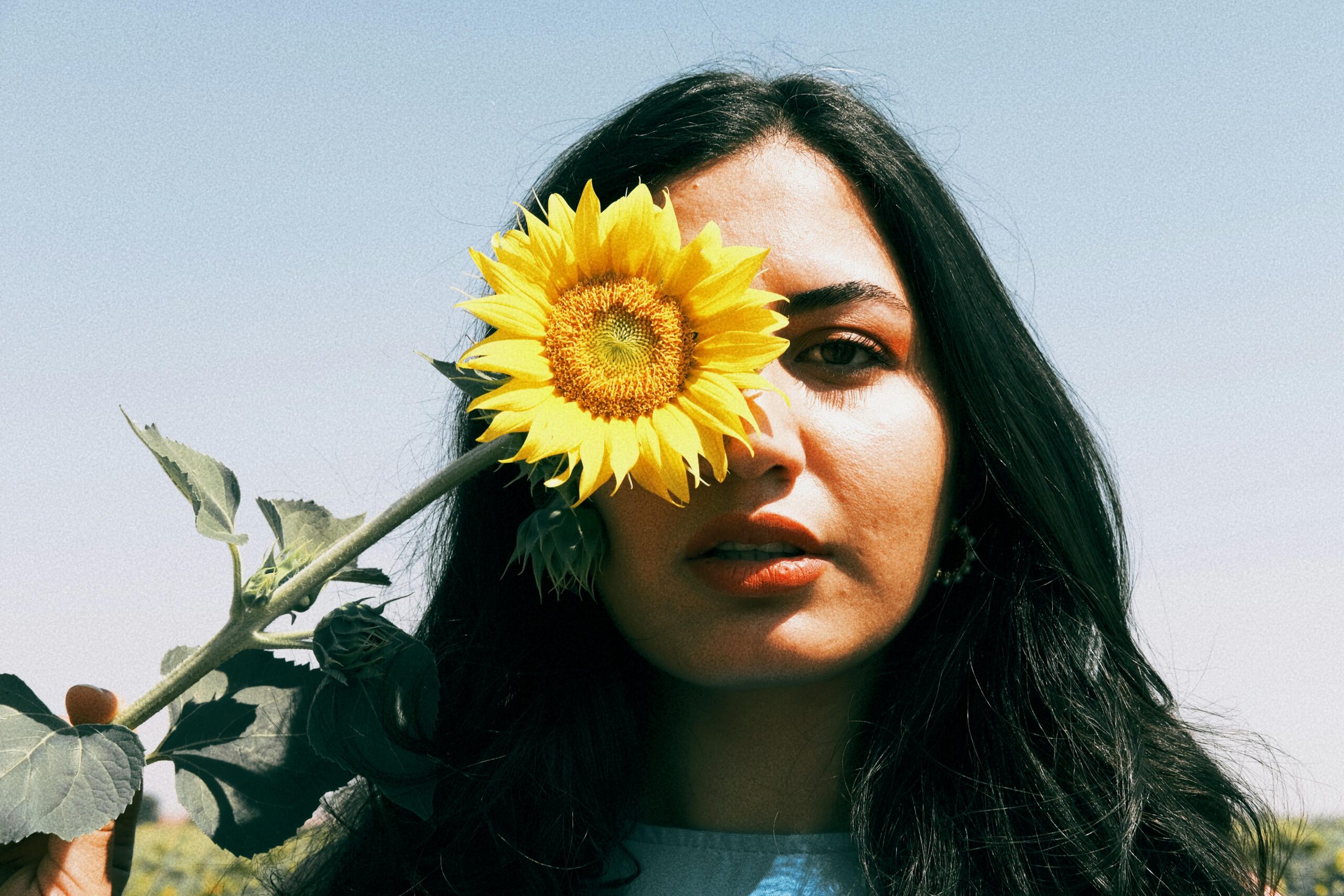 A woman with long dark hair stands outdoors against a clear sky, holding a bright yellow sunflower in front of one eye. The sunlight highlights her face as she looks directly at the camera, creating a serene and contemplative mood.