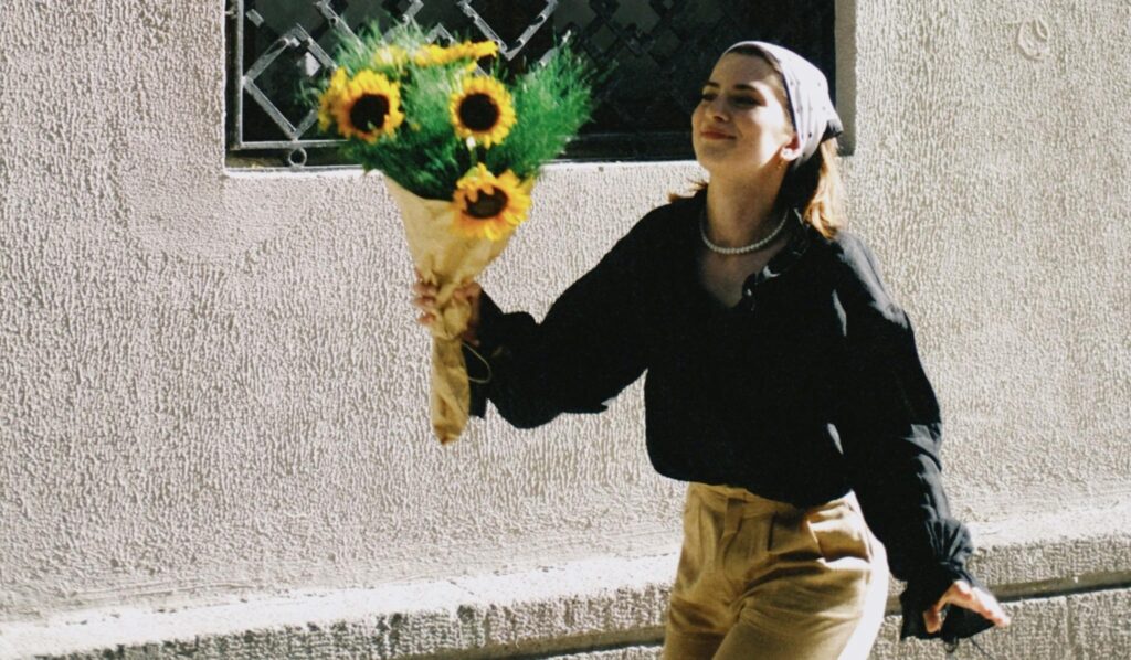 A woman walks happily down a sunlit street holding a bouquet of sunflowers wrapped in brown paper. She is wearing a black blouse, beige pants, a pearl necklace, and a light-colored headscarf, with a soft smile on her face. The textured wall behind her casts gentle shadows, creating a calm, nostalgic mood.
