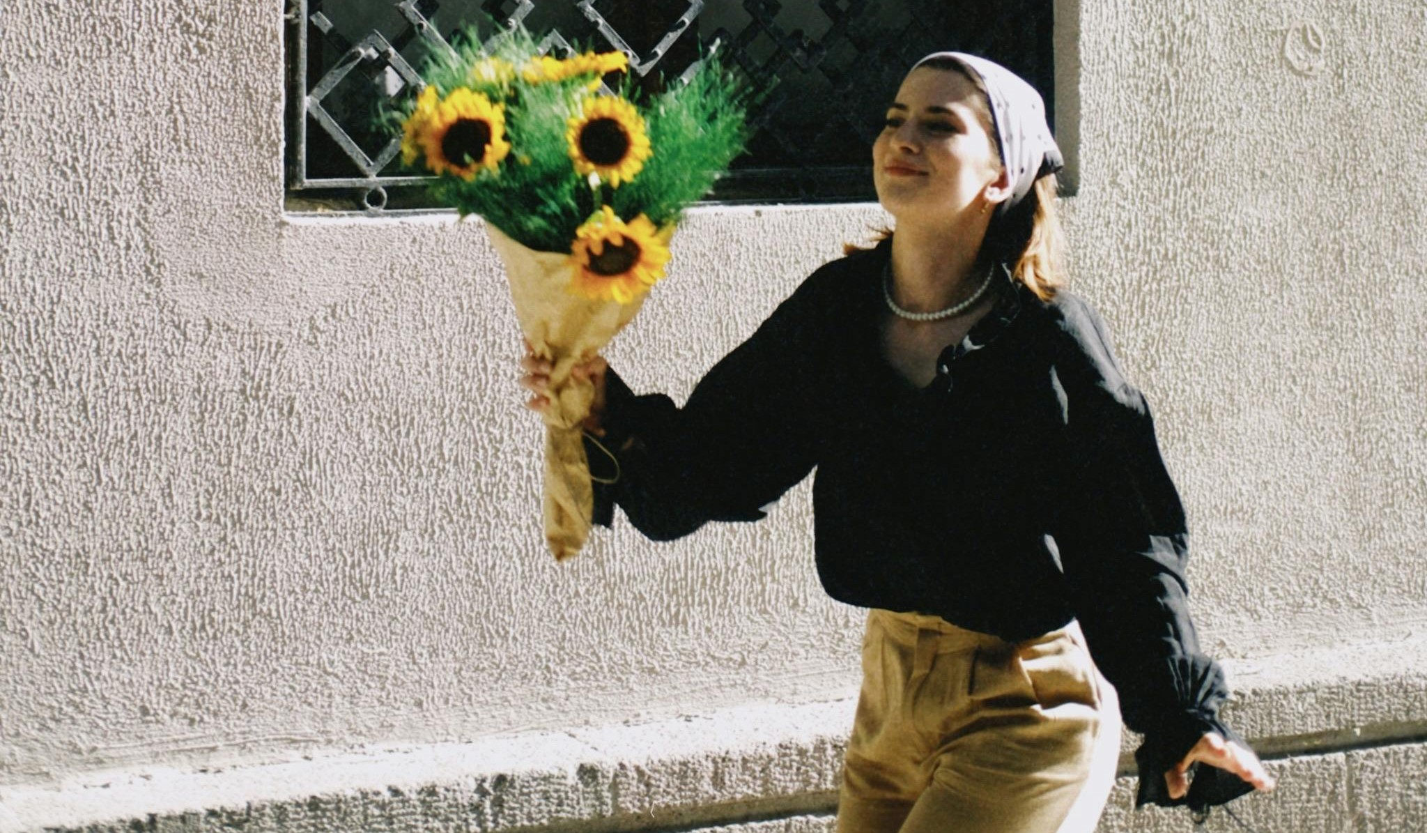 A woman walks happily down a sunlit street holding a bouquet of sunflowers wrapped in brown paper. She is wearing a black blouse, beige pants, a pearl necklace, and a light-colored headscarf, with a soft smile on her face. The textured wall behind her casts gentle shadows, creating a calm, nostalgic mood.