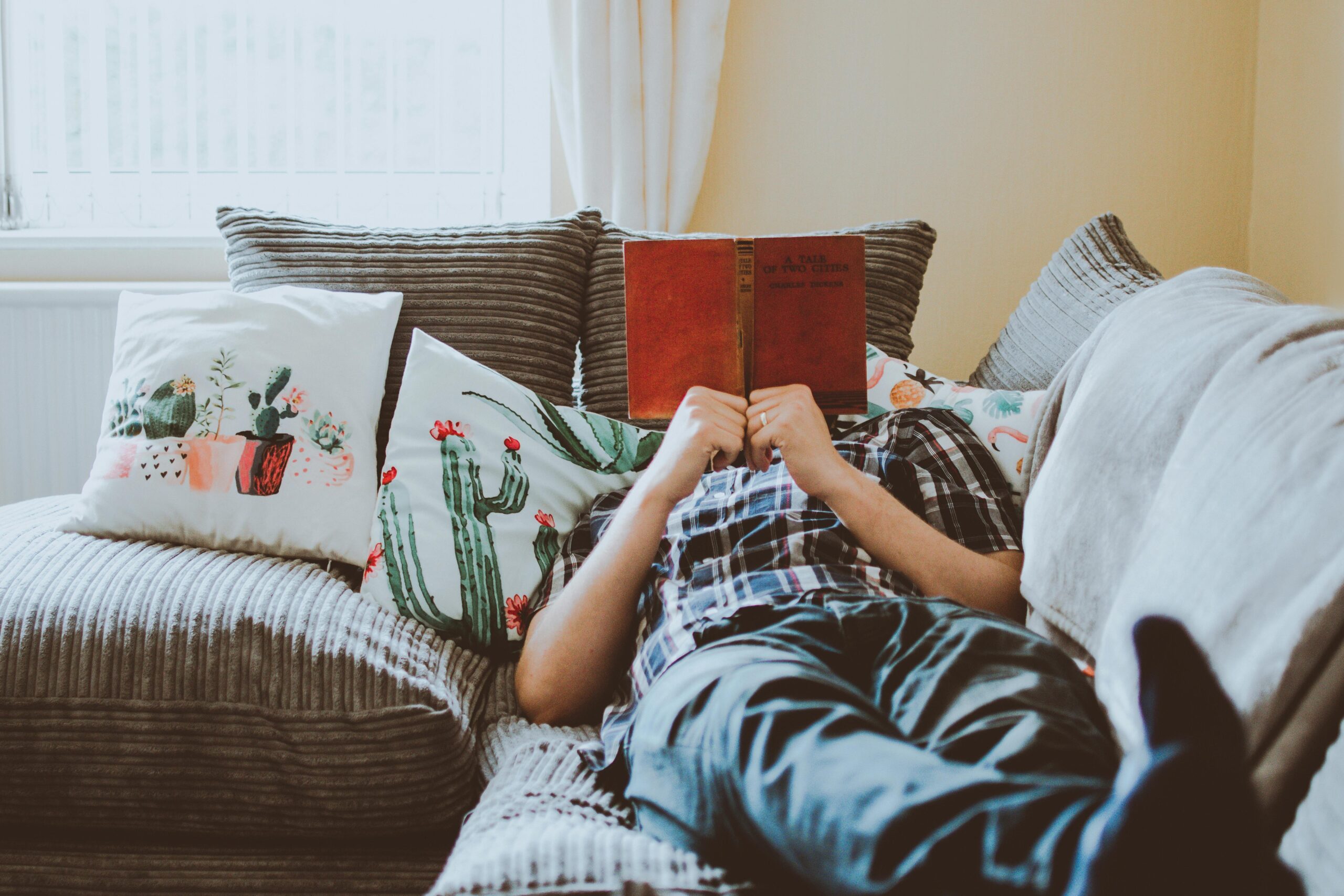Person relaxing on a sofa reading a book, surrounded by cozy patterned pillows and soft natural light — promoting rest and quiet moments.