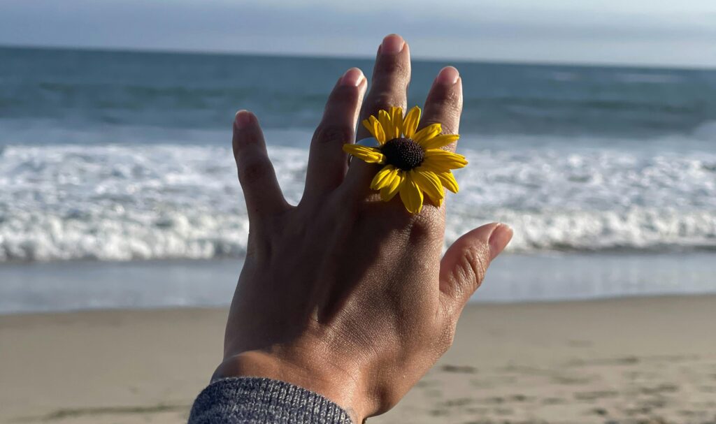 A hand with a yellow daisy ring held up toward the ocean waves, symbolizing mindfulness, renewal, and emotional balance.