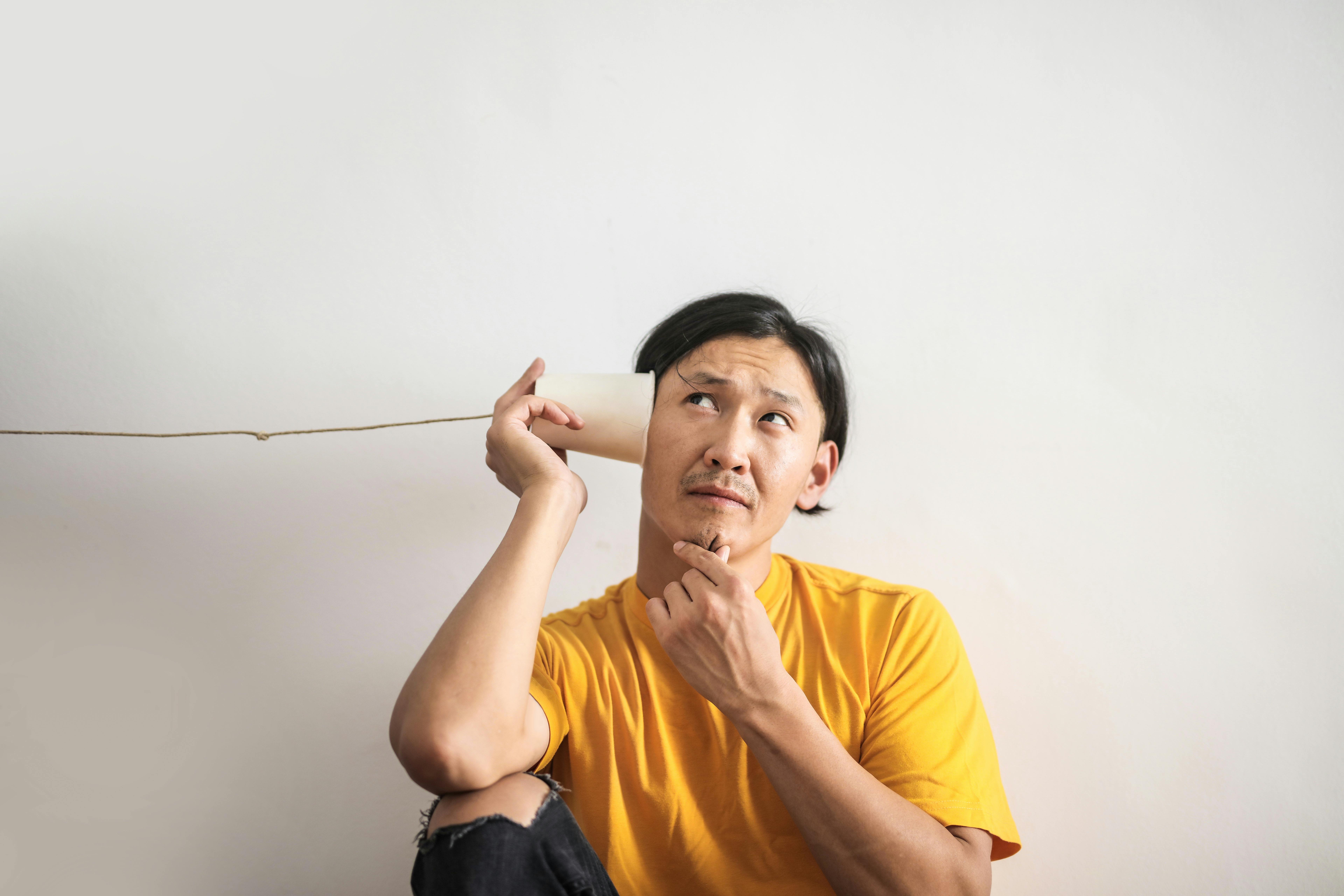A man wearing a yellow shirt sits thoughtfully while holding a paper cup phone to his ear, as if trying to listen through it. He looks upward in curiosity against a plain white wall.