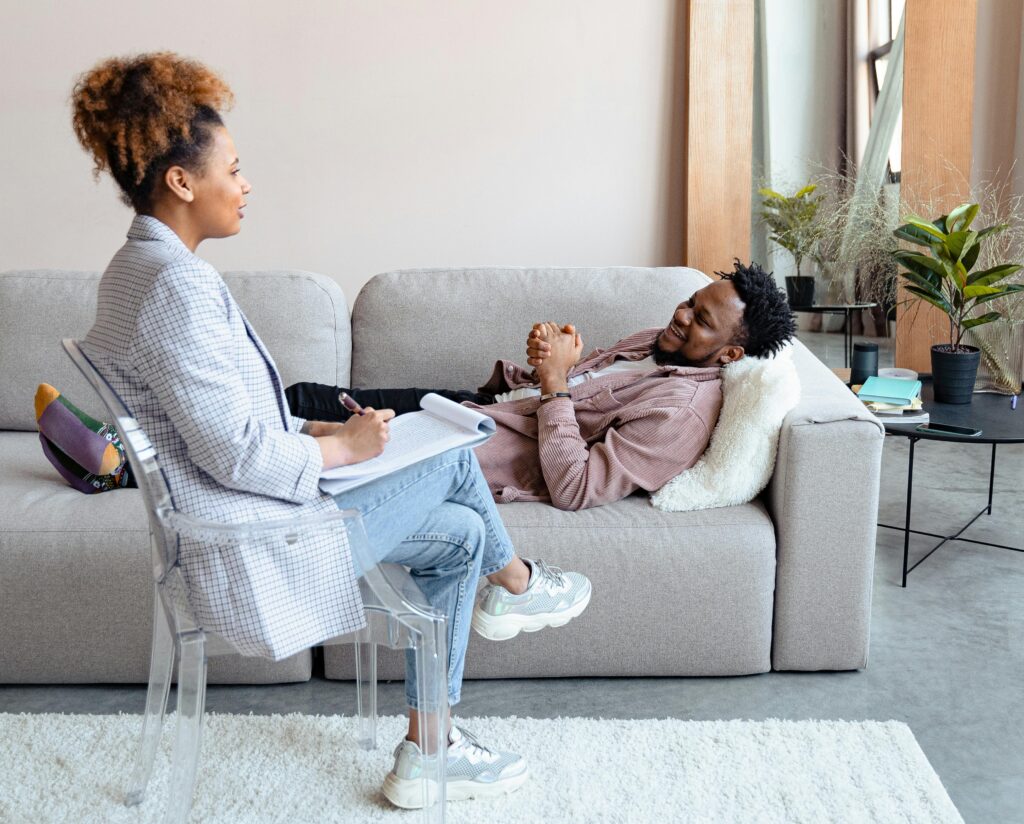 A therapist sits with a notebook while listening to a smiling client lying comfortably on a couch during a counseling session in a warm, modern therapy office.