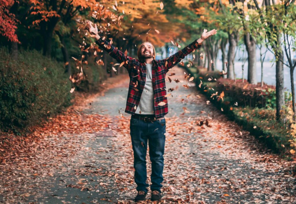 A joyful person standing on a tree-lined path, tossing autumn leaves into the air, celebrating the beauty of fall and mindfulness in every season.