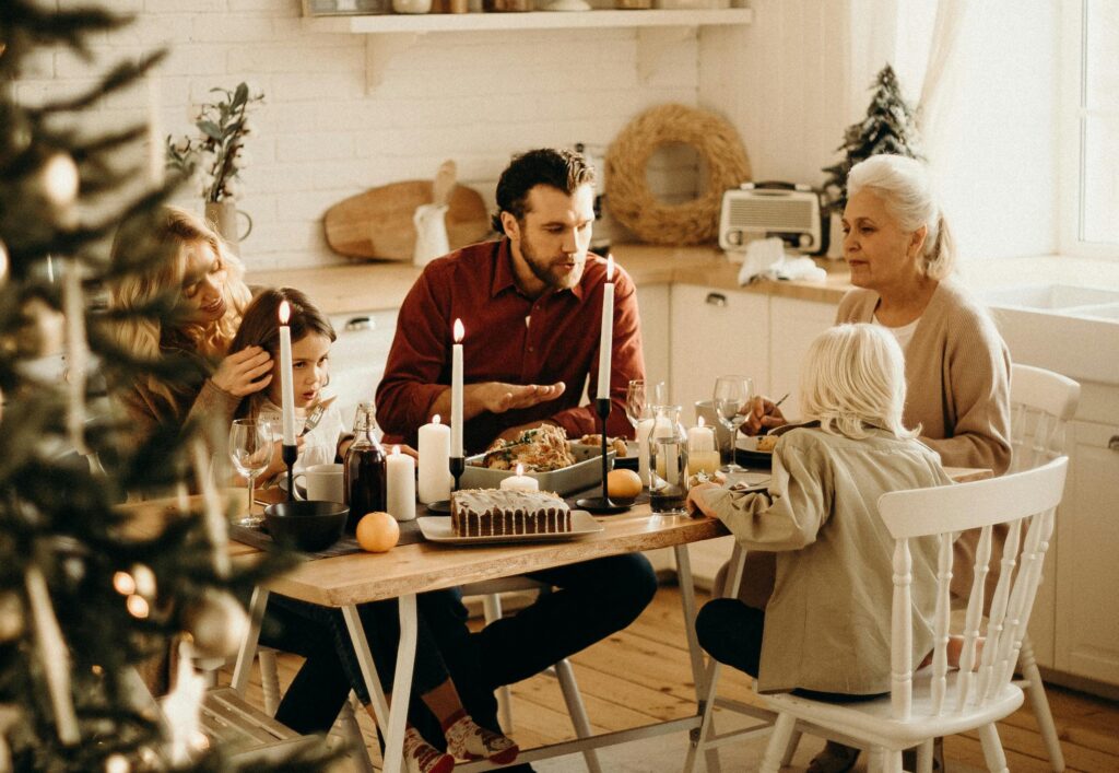 A multigenerational family sits around a candlelit dining table during a holiday meal, with adults and children engaged in quiet conversation in a warm, softly lit kitchen decorated for the season.