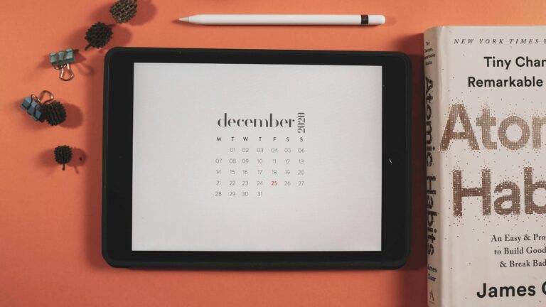 Warm, minimalist desk scene with a December calendar on a tablet and Atomic Habits nearby, evoking a season of pause, reflection, and reset.”