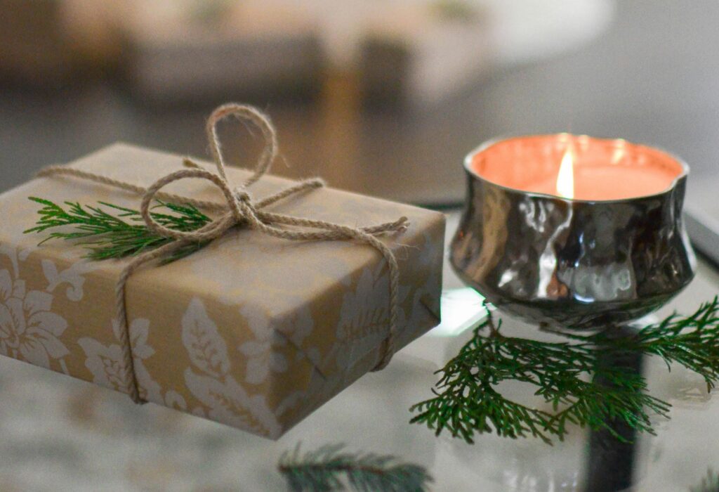 A softly wrapped gift tied with twine sits beside a lit candle and evergreen sprigs, creating a calm, cozy scene that evokes peace, rest, and intentional slowing down.