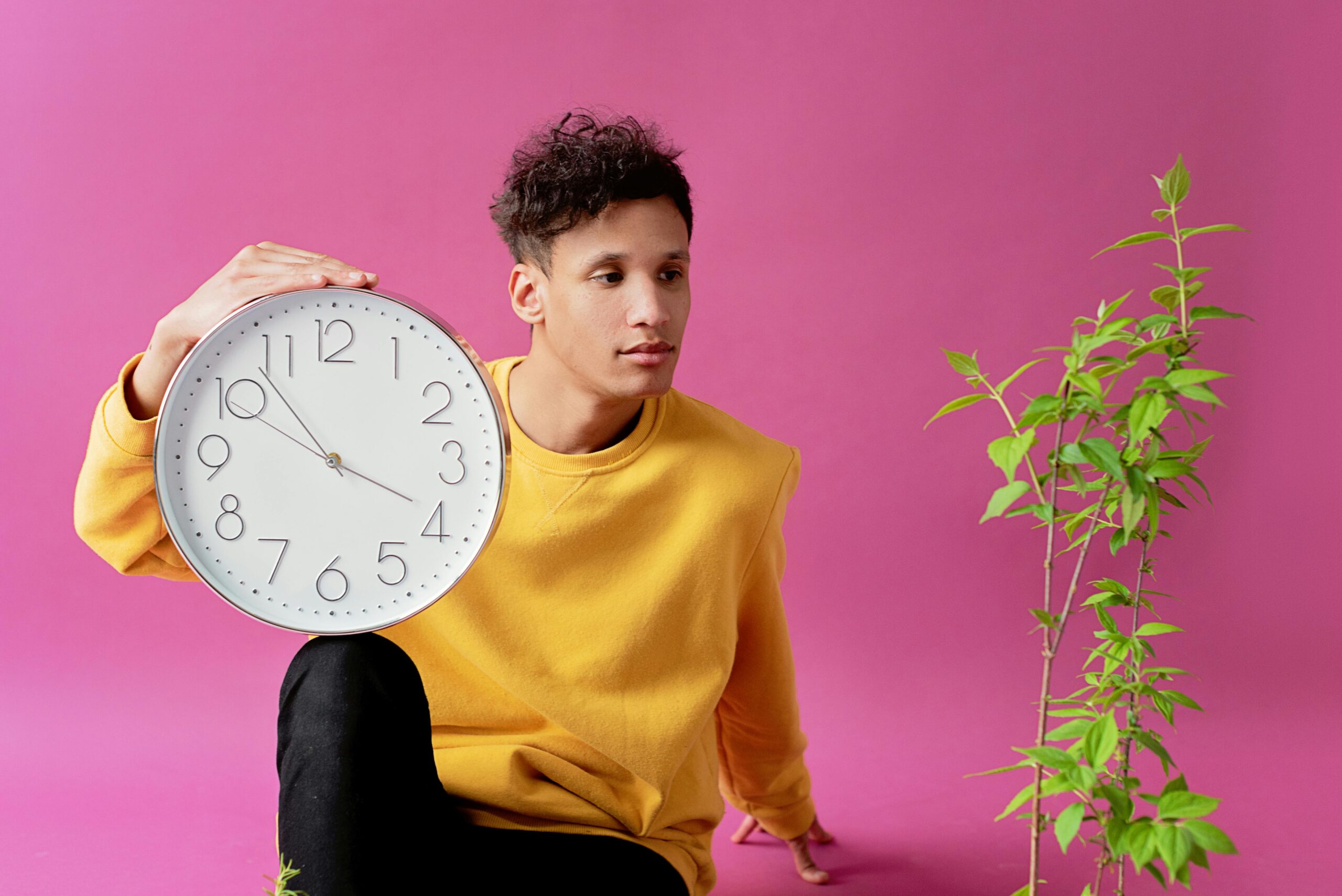 A person in a yellow sweater sits against a pink background, holding a large wall clock beside them while looking calmly to the side, with a small green plant nearby—suggesting patience, growth, and moving at one’s own pace.