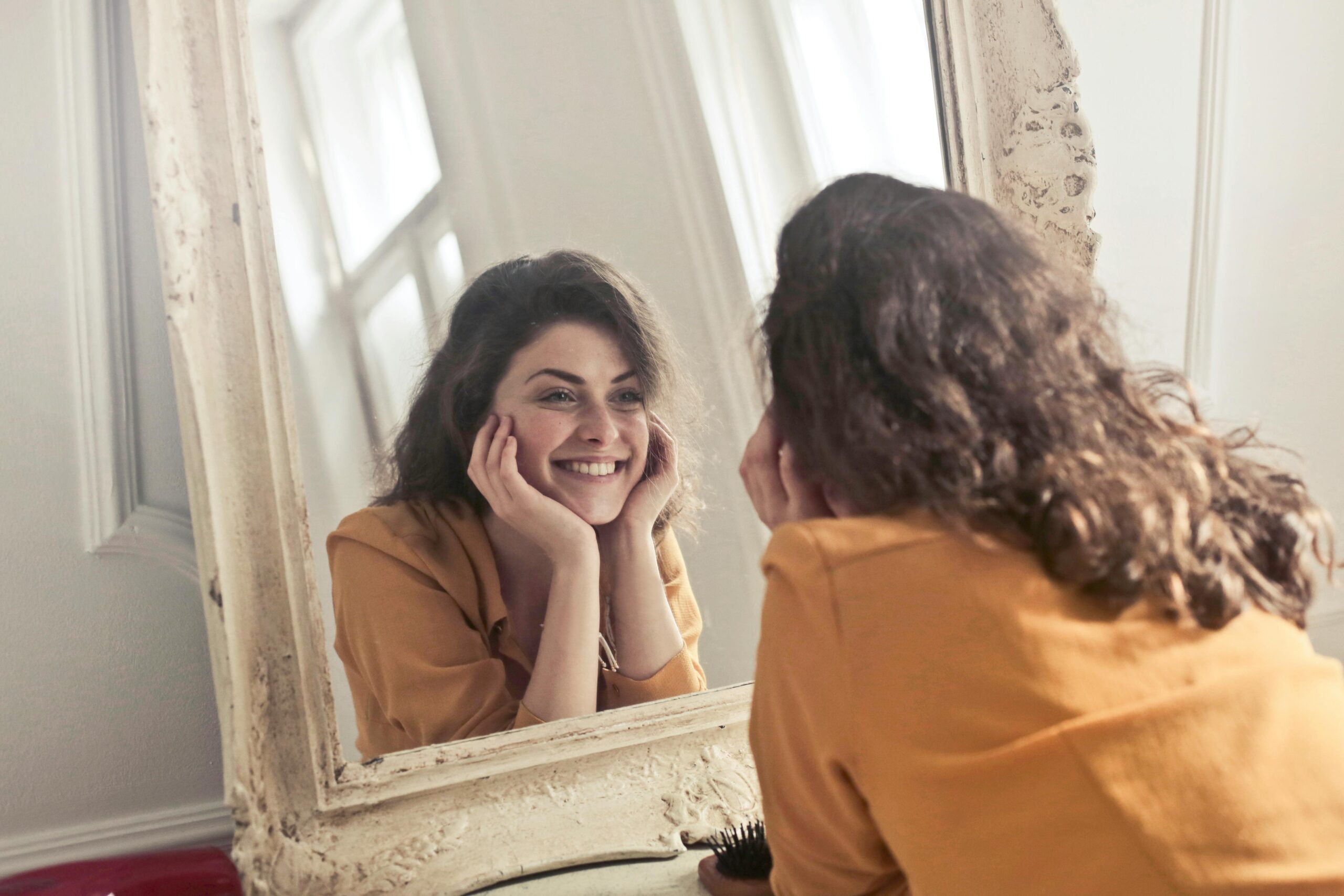 A woman in a mustard-colored top smiles warmly at her reflection in a large antique mirror, resting her face in her hands in a bright, softly lit room.