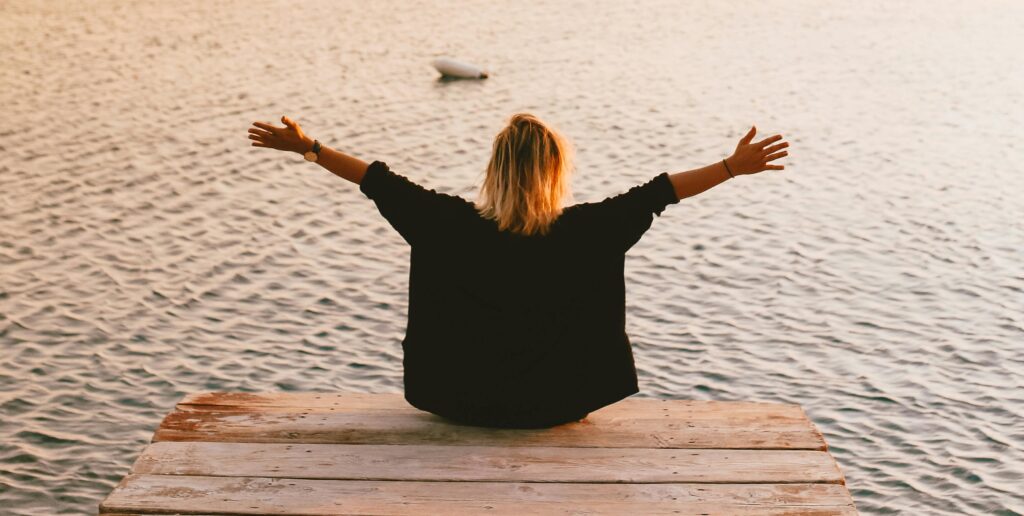 A person with blond hair sits on the edge of a wooden dock with arms outstretched, facing a calm body of water at sunset, conveying a sense of peace and openness.