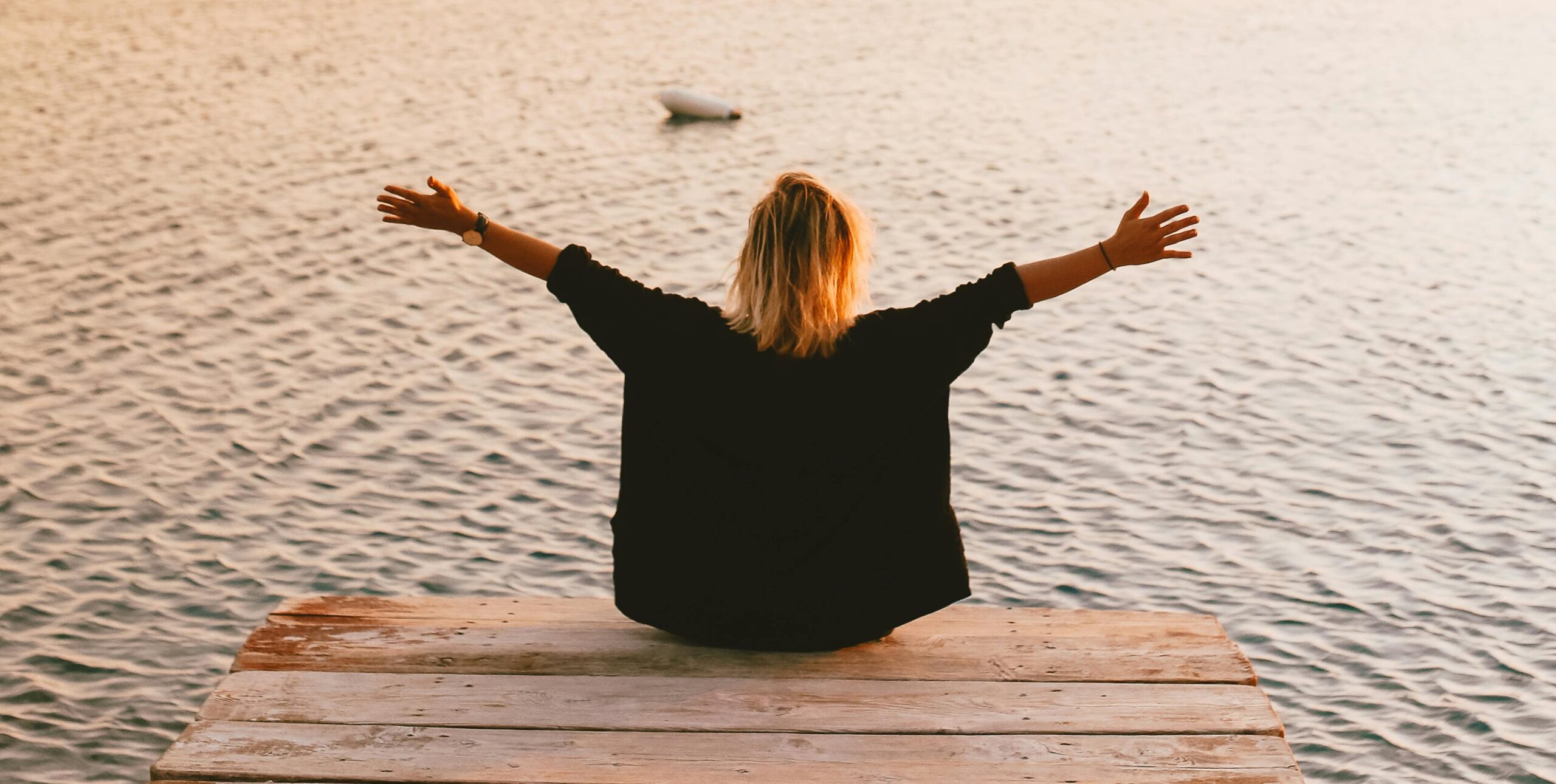 A person with blond hair sits on the edge of a wooden dock with arms outstretched, facing a calm body of water at sunset, conveying a sense of peace and openness.