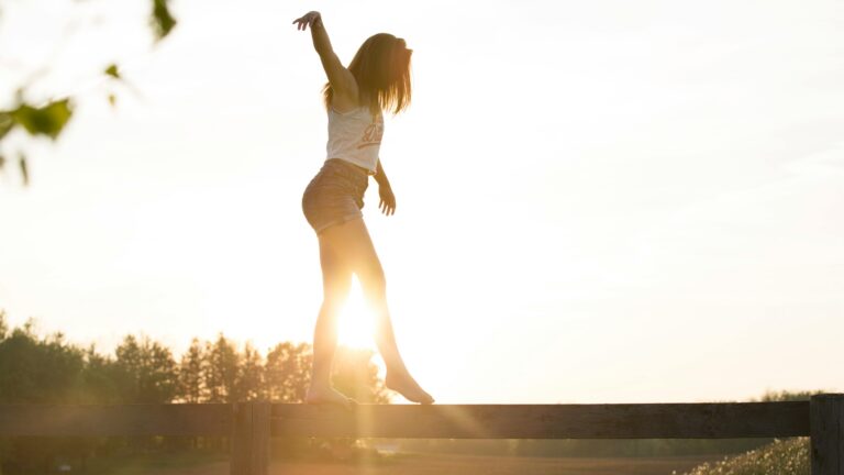 A woman balances barefoot on a wooden fence at sunset, her arms gently outstretched as warm sunlight glows behind her. Trees and open fields sit softly blurred in the background.