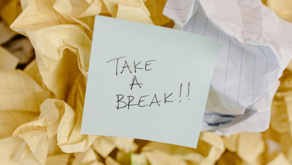 A handwritten sticky note reading “Take a Break!!” sits on top of a pile of crumpled lined paper, suggesting stress and the need for rest.