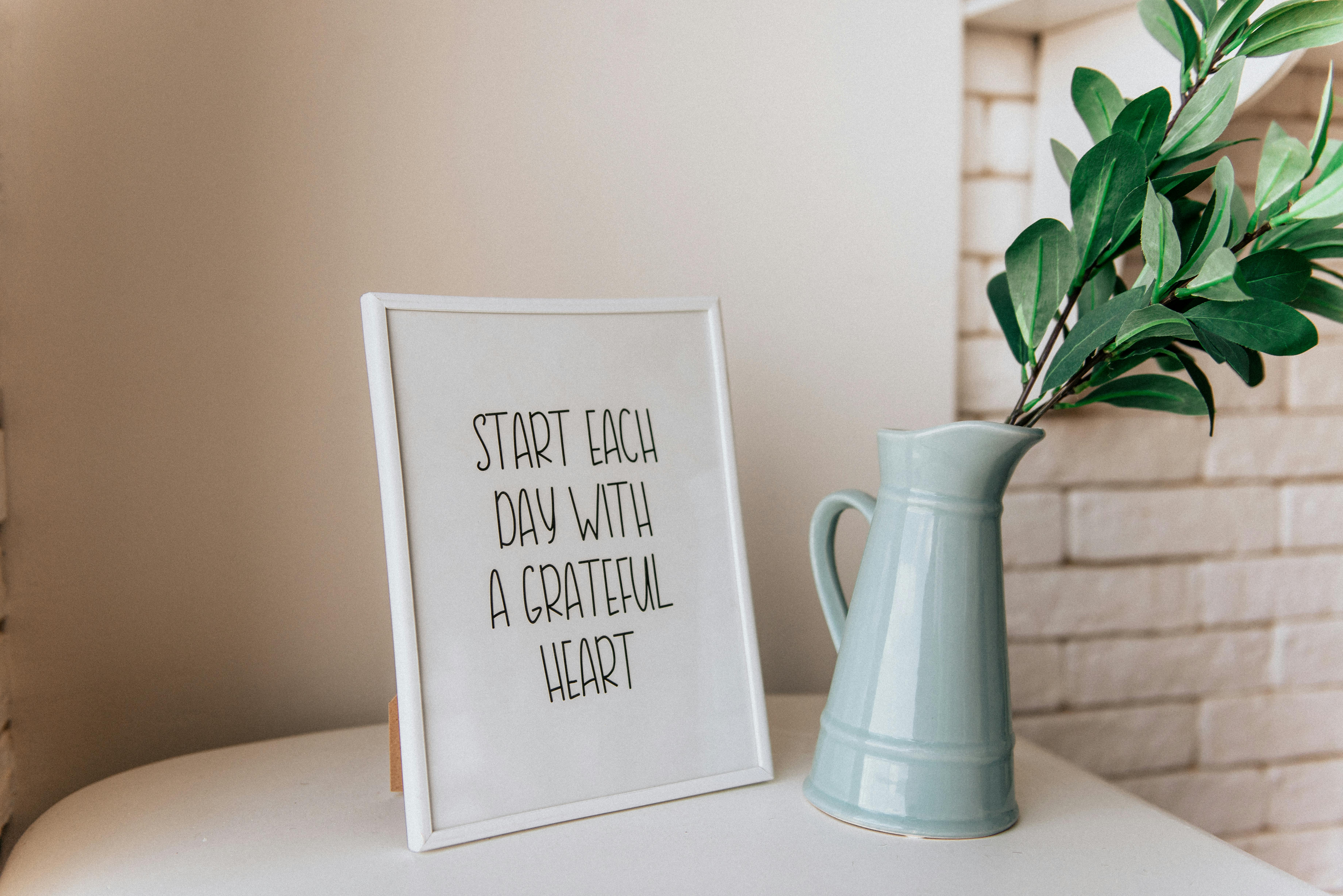 Framed quote reading ‘Start each day with a grateful heart’ displayed on a white table next to a pale blue vase with greenery, creating a peaceful, reflective mood.