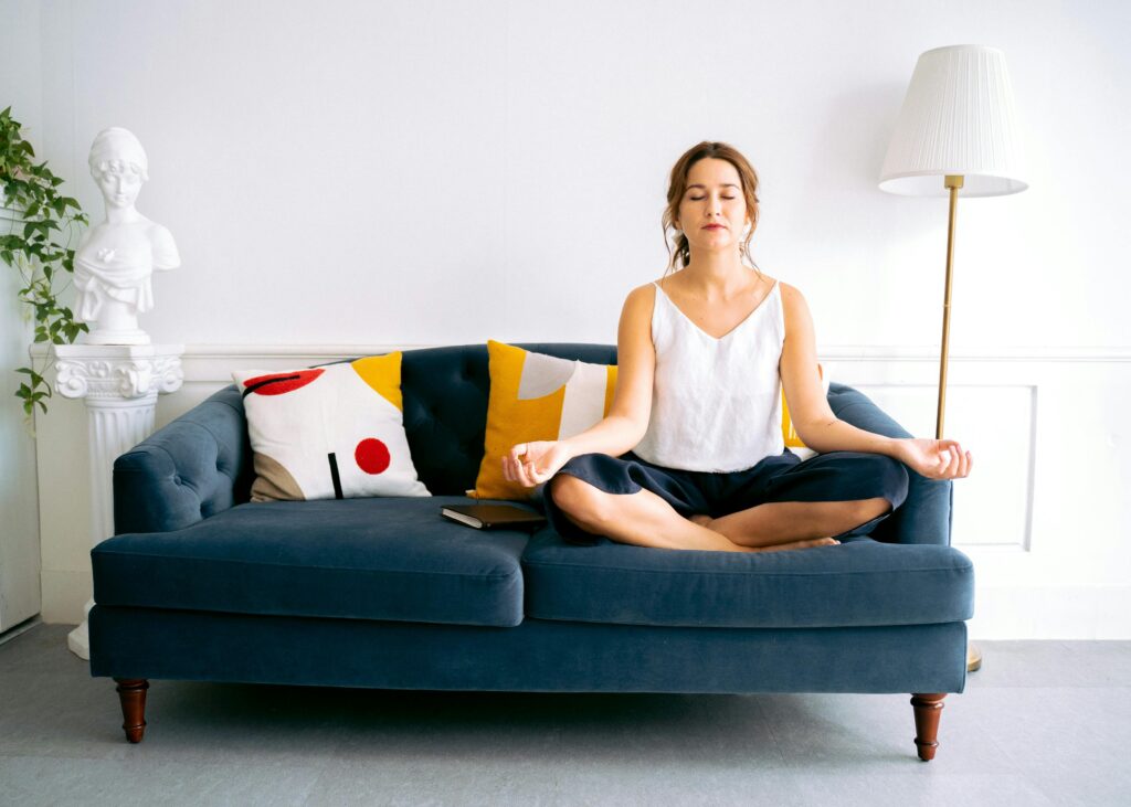 Woman sits cross-legged on a blue sofa with eyes closed, meditating in a bright, minimalist living room.