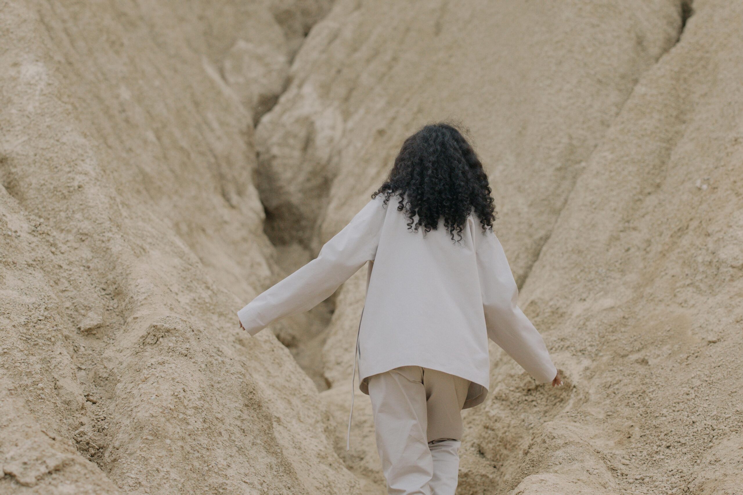 Person with curly hair, seen from behind, walking through a narrow sandy rock canyon with arms slightly outstretched.