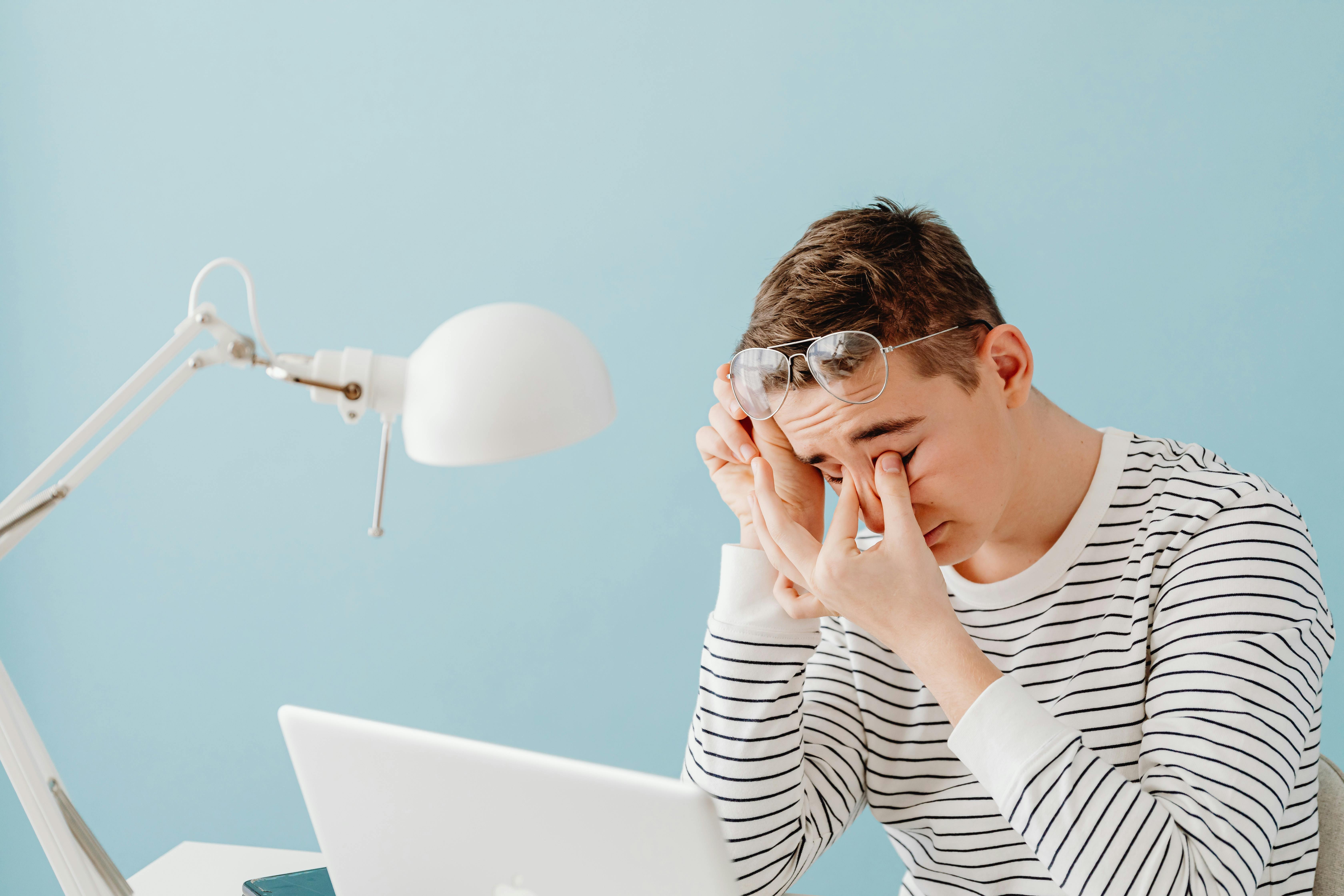Person wearing a striped shirt sits at a desk rubbing their eyes, glasses pushed up on their forehead, looking fatigued while working on a laptop against a light blue background.