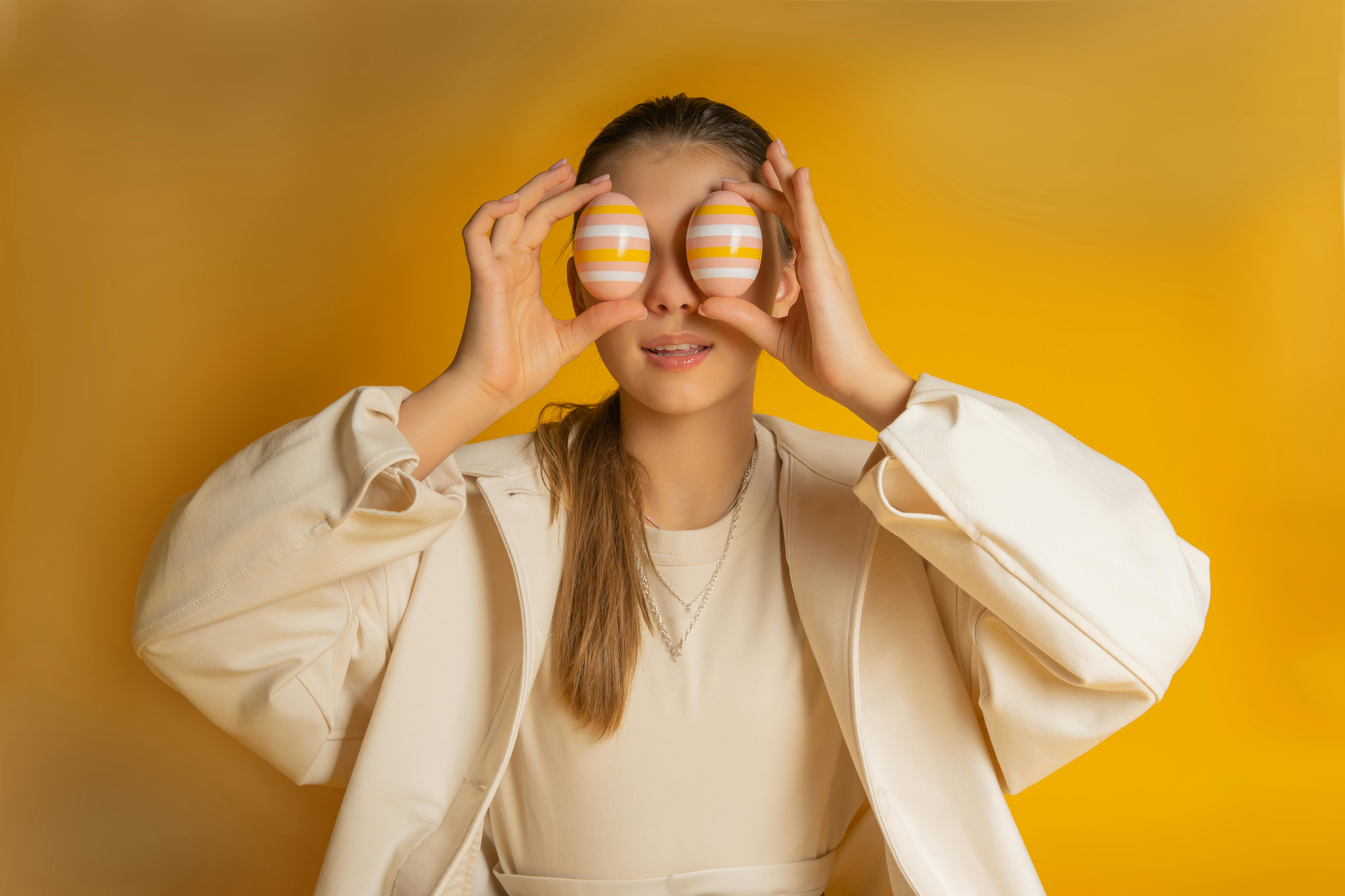 Woman holding two striped pastel eggs over her eyes against a bright yellow background, playfully covering her vision for a “5 Things You Can See” anxiety grounding exercise theme.