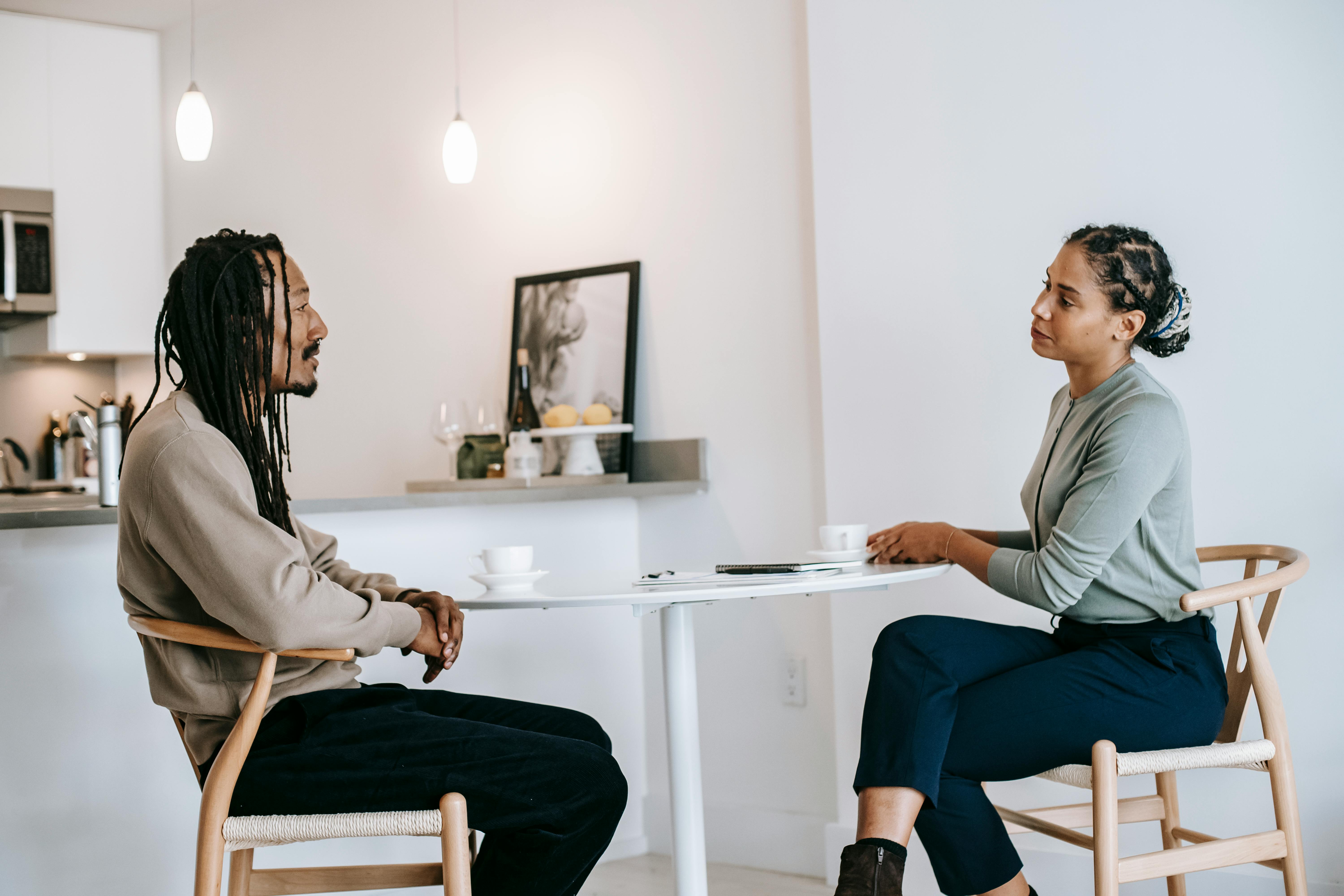 Two adults seated across from each other at a small table in a bright, minimalist room, engaged in a calm, focused conversation resembling a therapy session, with notebooks and coffee cups on the table.