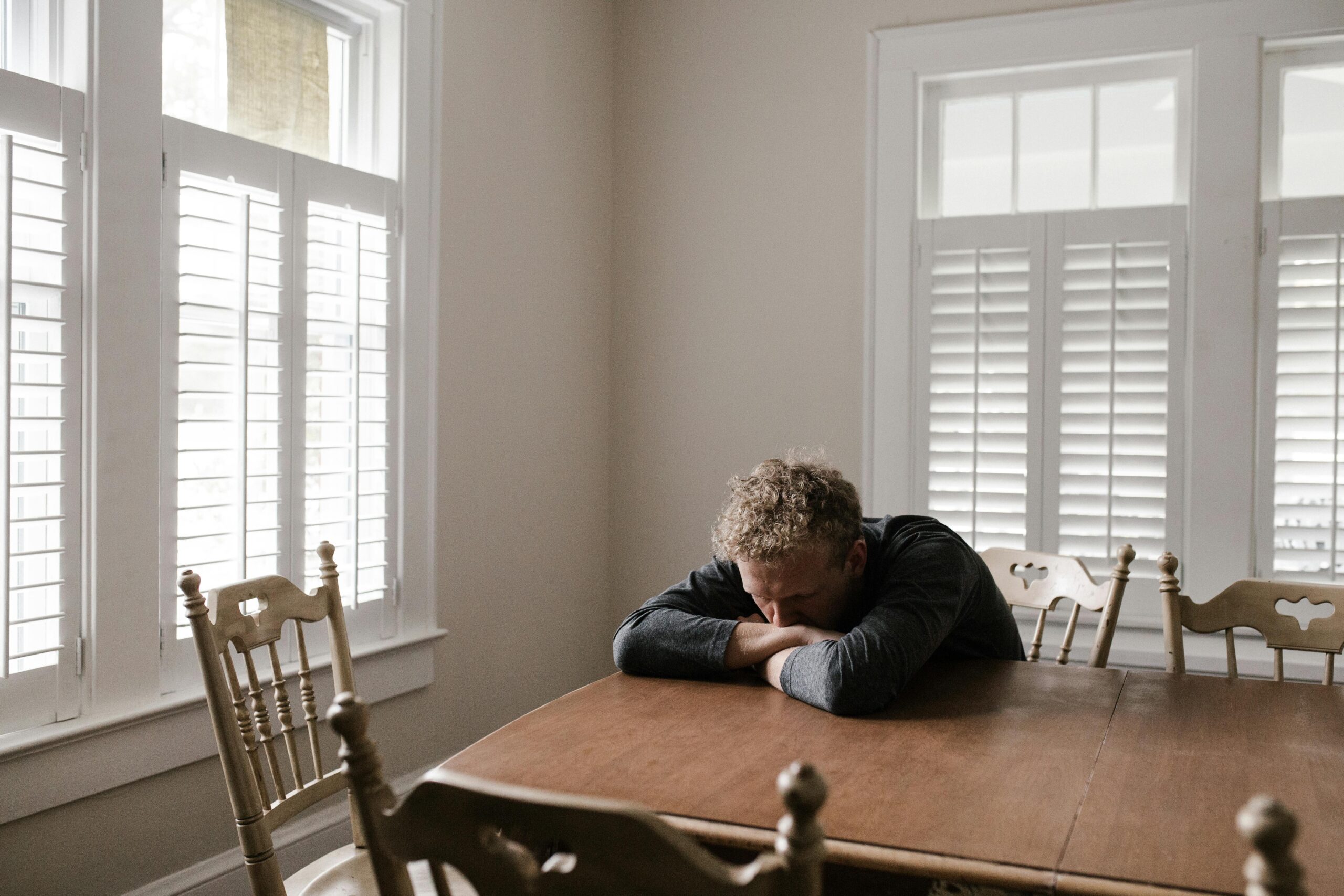 A man sits alone at a wooden dining table with his head resting on his folded arms, surrounded by empty chairs and soft daylight coming through shuttered windows, conveying exhaustion and emotional stress.