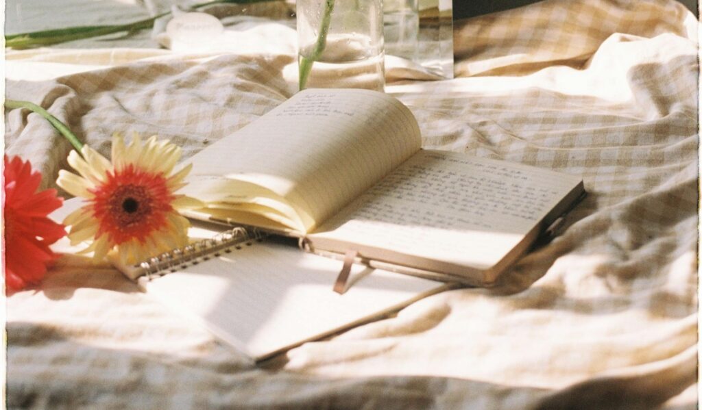 An open journal with handwritten notes rests on a sunlit bed beside a small notebook and bright gerbera daisies, creating a calm, reflective mood-tracking scene.