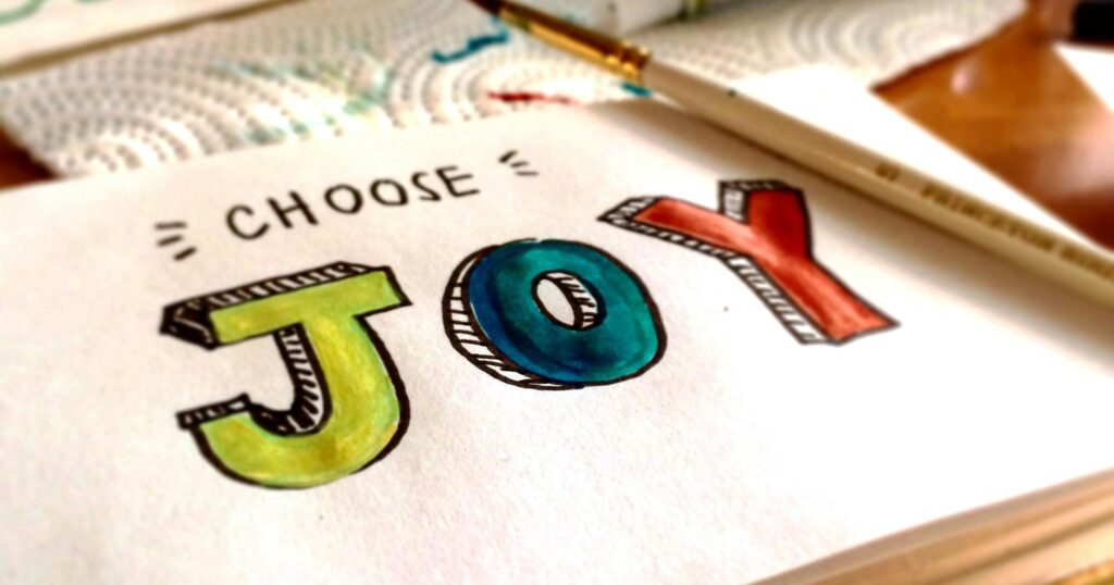 Hand-drawn, colorful lettering spelling “Choose Joy” on white paper, with a paintbrush resting nearby, symbolizing creativity, positivity, and making joy a daily choice.