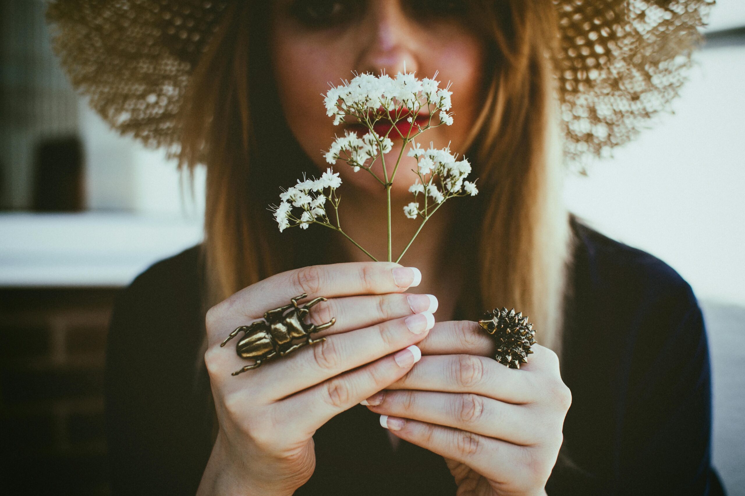 Person wearing a wide-brim hat holds delicate white flowers up to their face, with decorative statement rings visible on their fingers, conveying a calm and reflective mood.
